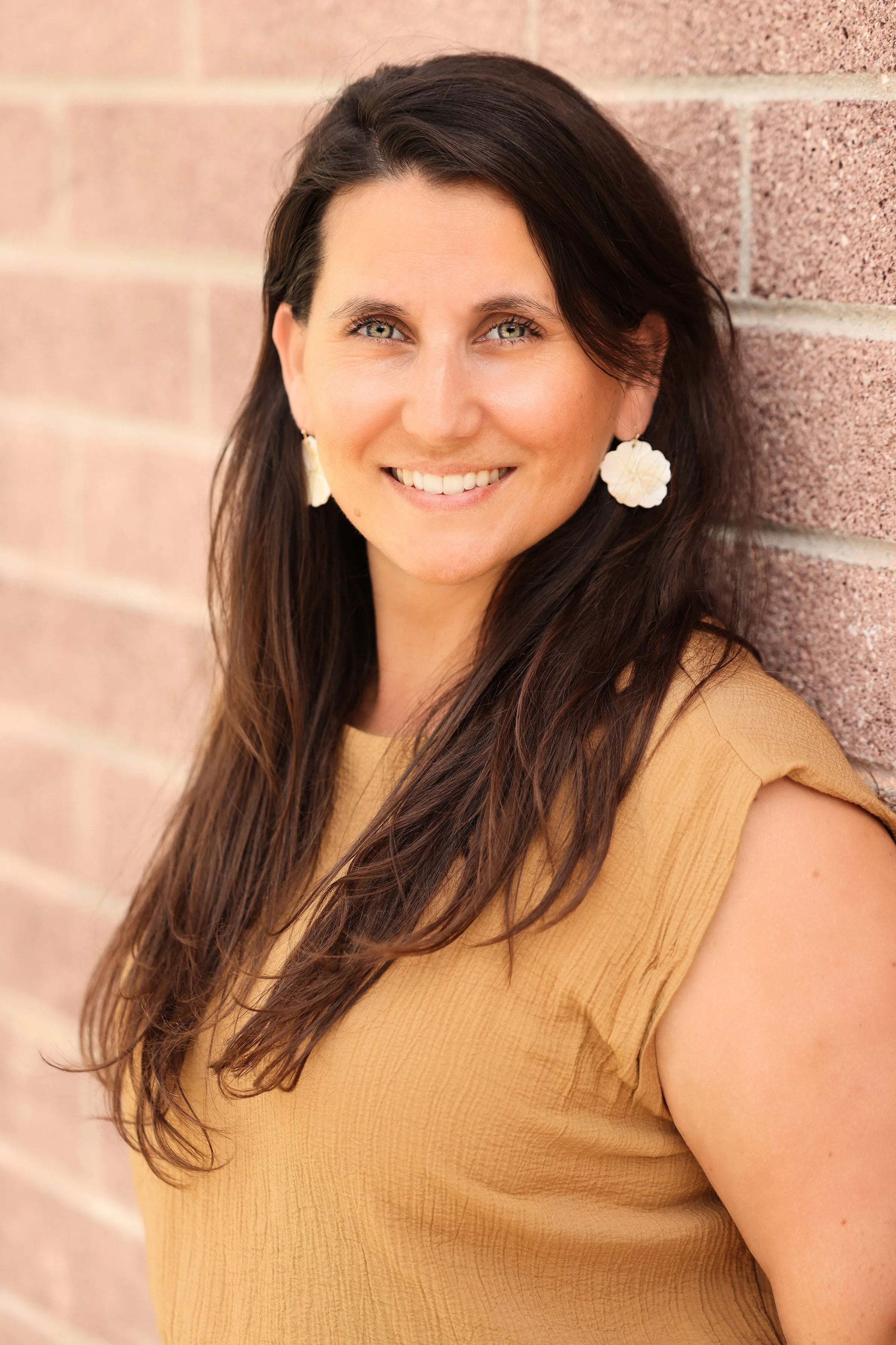 A woman with long dark hair, smiling, leaning against a brick wall, wearing a sleeveless beige top and white floral earrings.