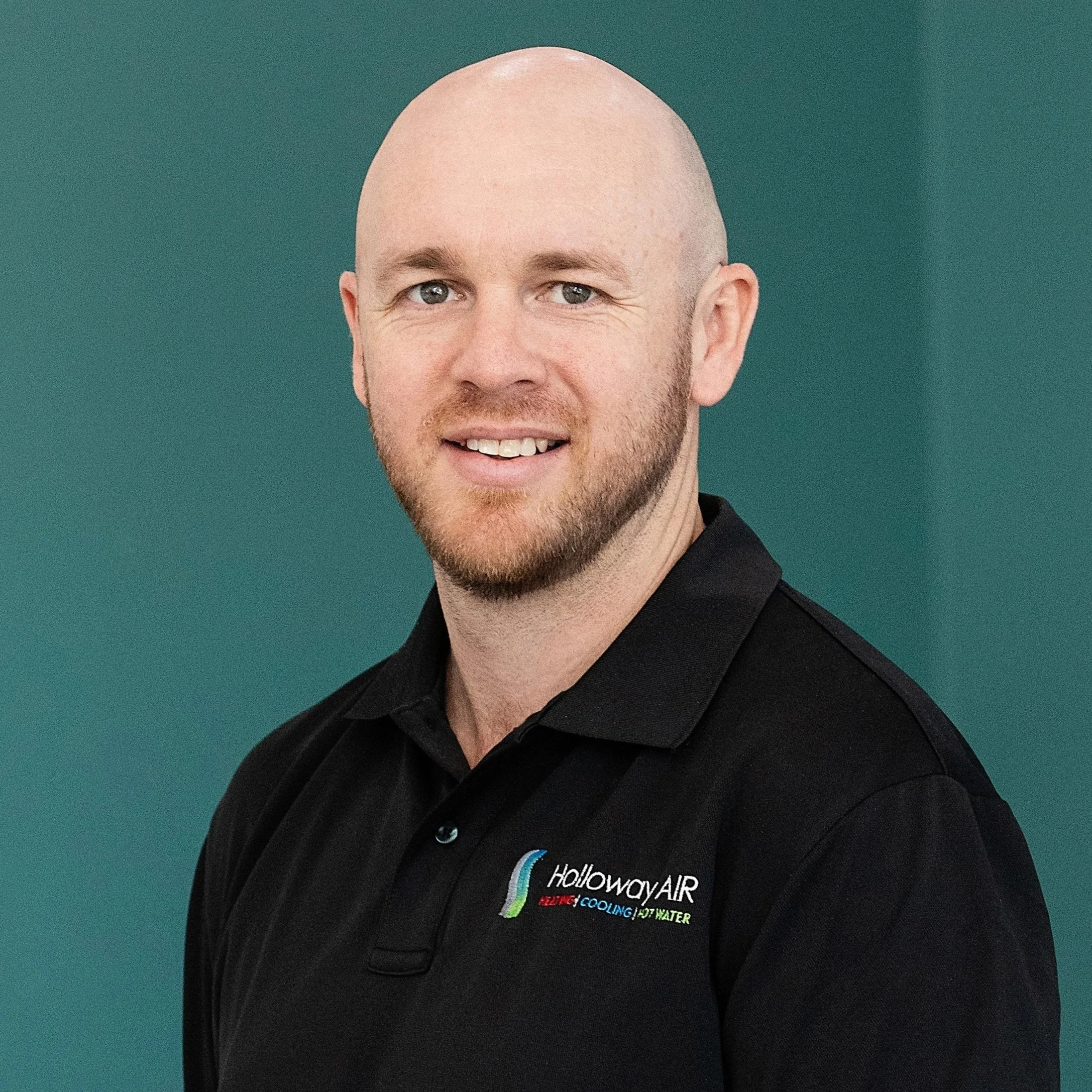 A man with a bald head, light beard, and blue eyes smiling at the camera. He is wearing a black polo shirt with a colored logo and text that reads 'HollowayAIR' and related services on it. The background is a solid greenish color.