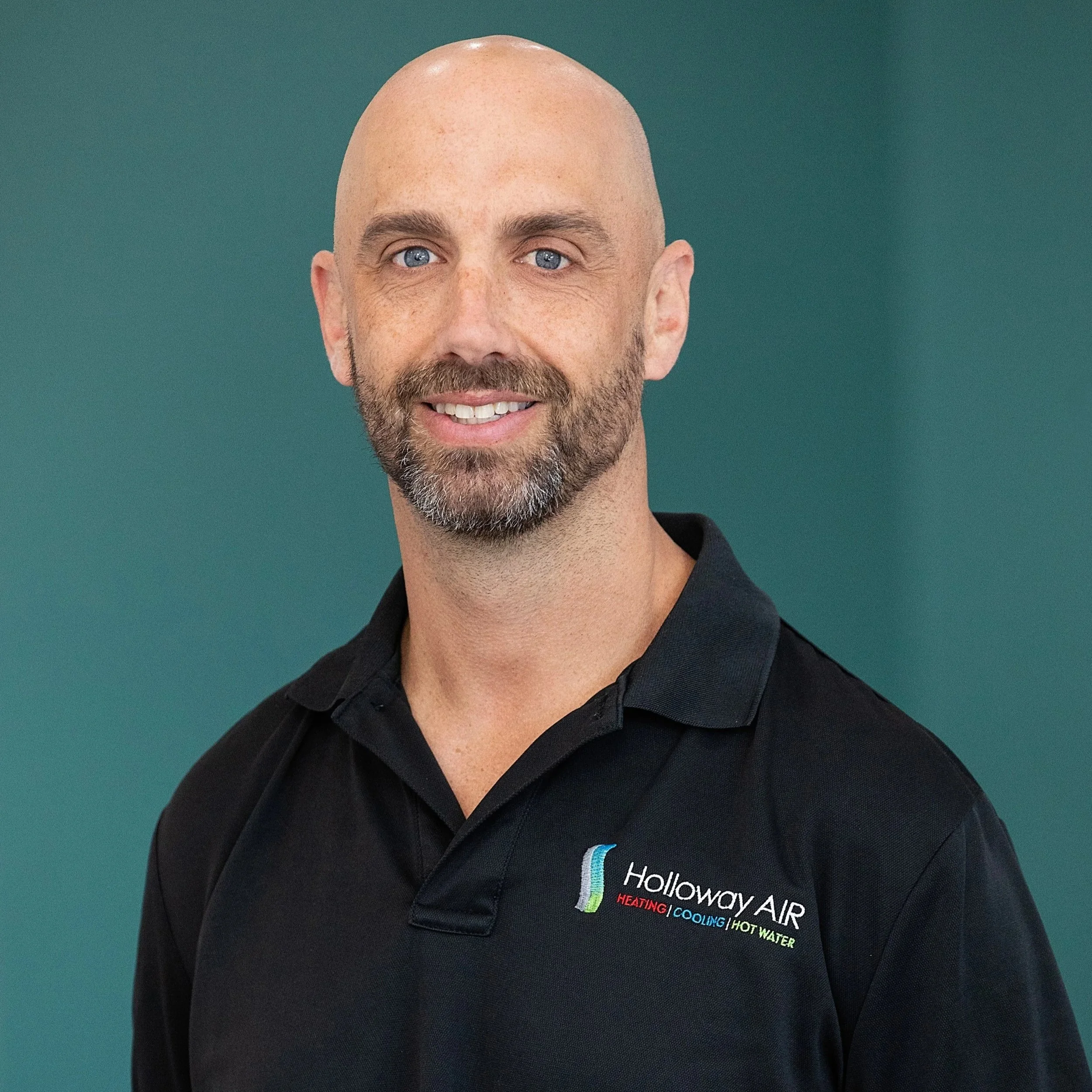 A man with a beard and bald head smiling, wearing a black polo shirt with "Holloway AIR" logo and text about heating, cooling, and hot water, against a plain teal background.