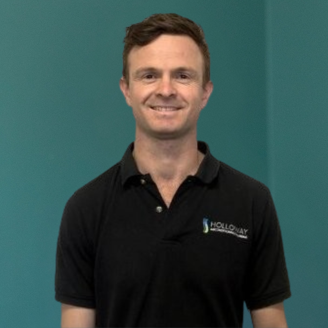 A man with short brown hair smiling, wearing a black polo shirt with a logo, standing against a teal background.