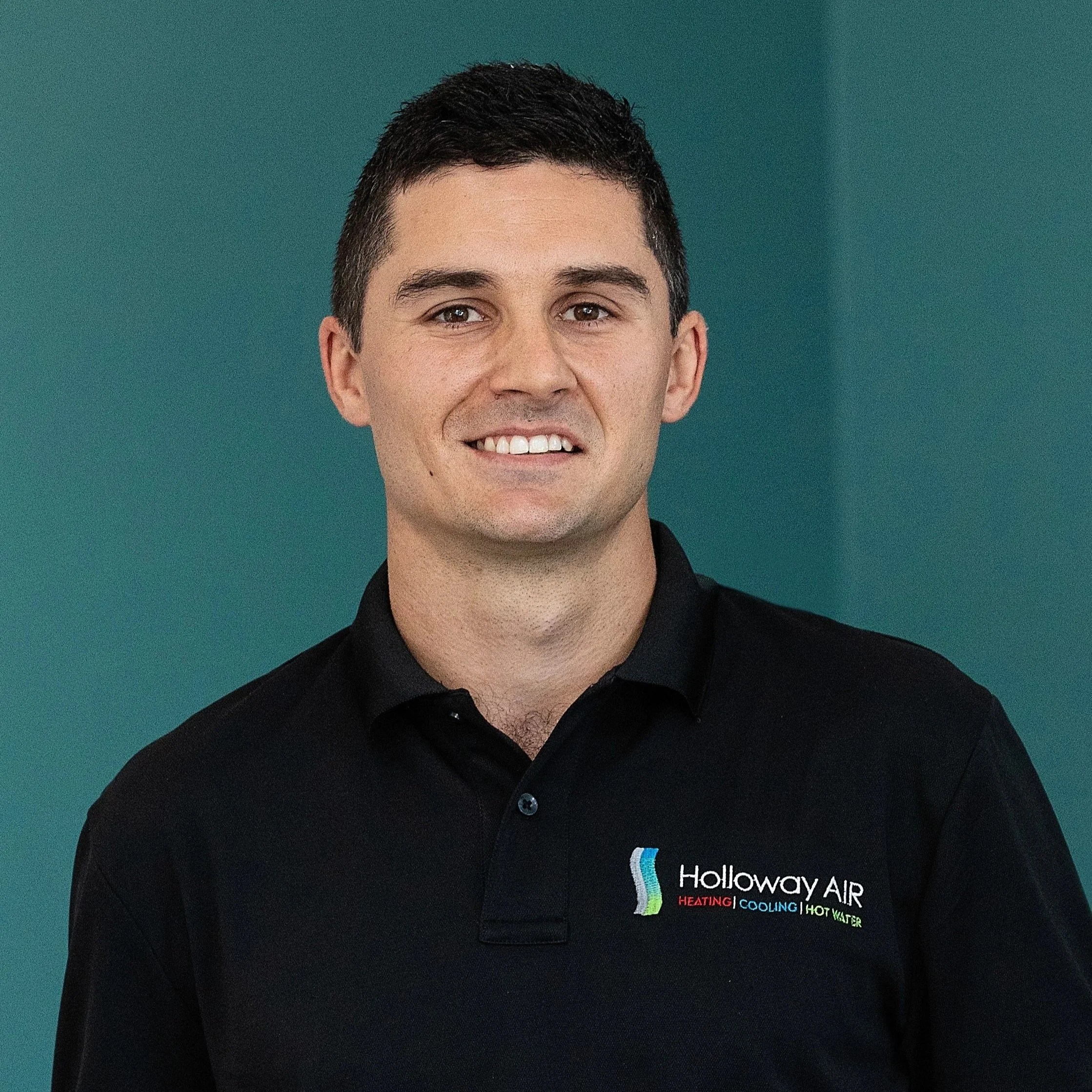 A young man with short dark hair wearing a black polo shirt with a logo for Holloway Air, which includes the words HEATING, COOLING, and HOT WATER, standing against a teal background.