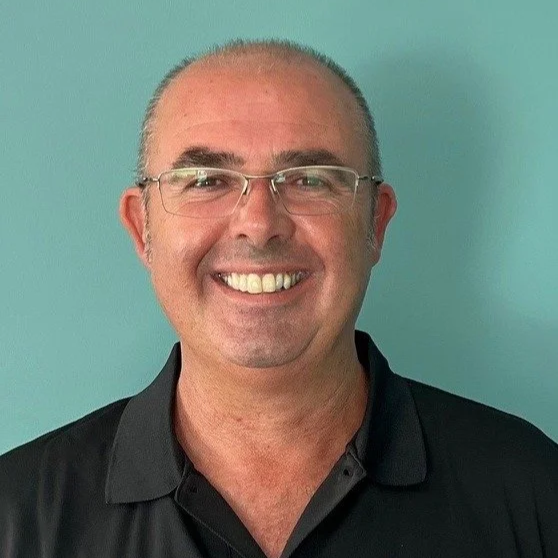 A man with glasses and short hair, smiling, wearing a black polo shirt, standing against a light teal background.