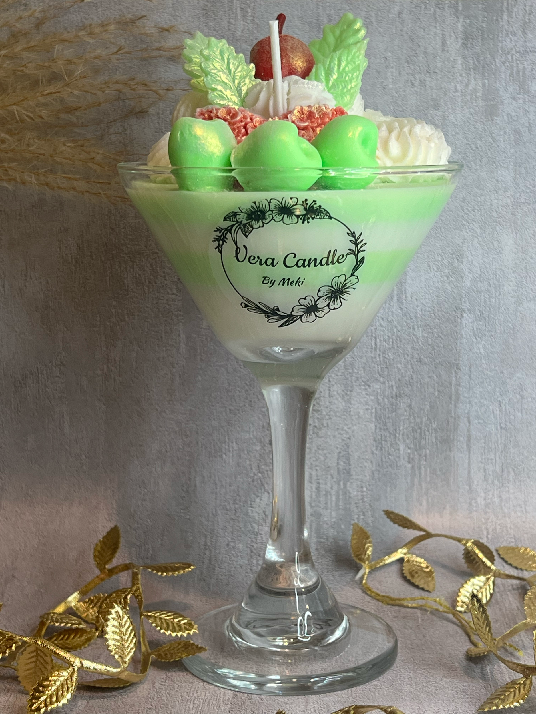 Decorative candle in a glass holder labeled "Vera Candle by Meki," designed to resemble a layered dessert with green and white wax, topped with green and red candy-like decorations, faux leaves, and a small candle, surrounded by gold metallic leaves.