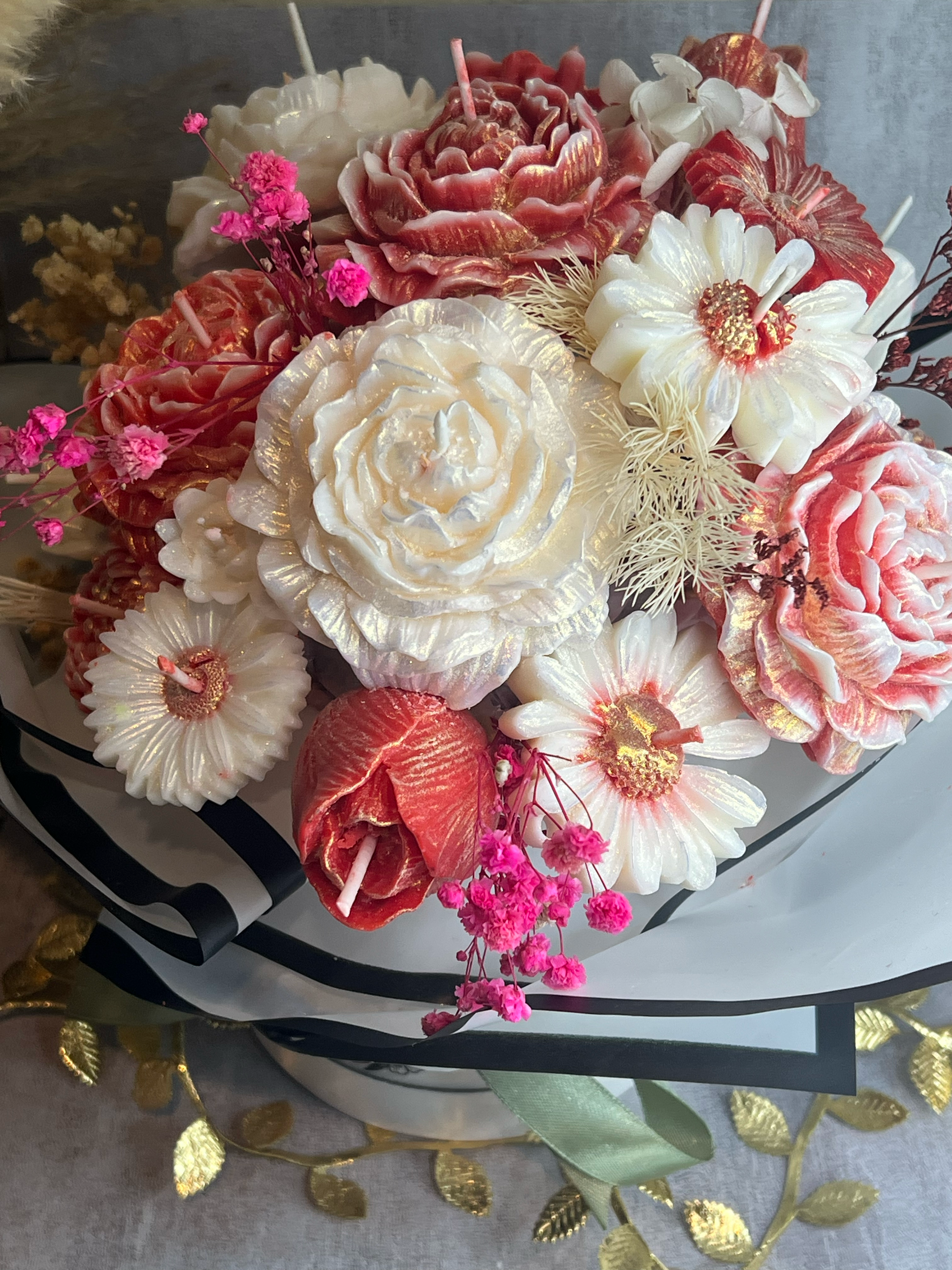 A decorative bouquet of candles shaped like flowers, including roses and daisies, with pink accents and small pink flowers, arranged in a black and white striped wrapping.