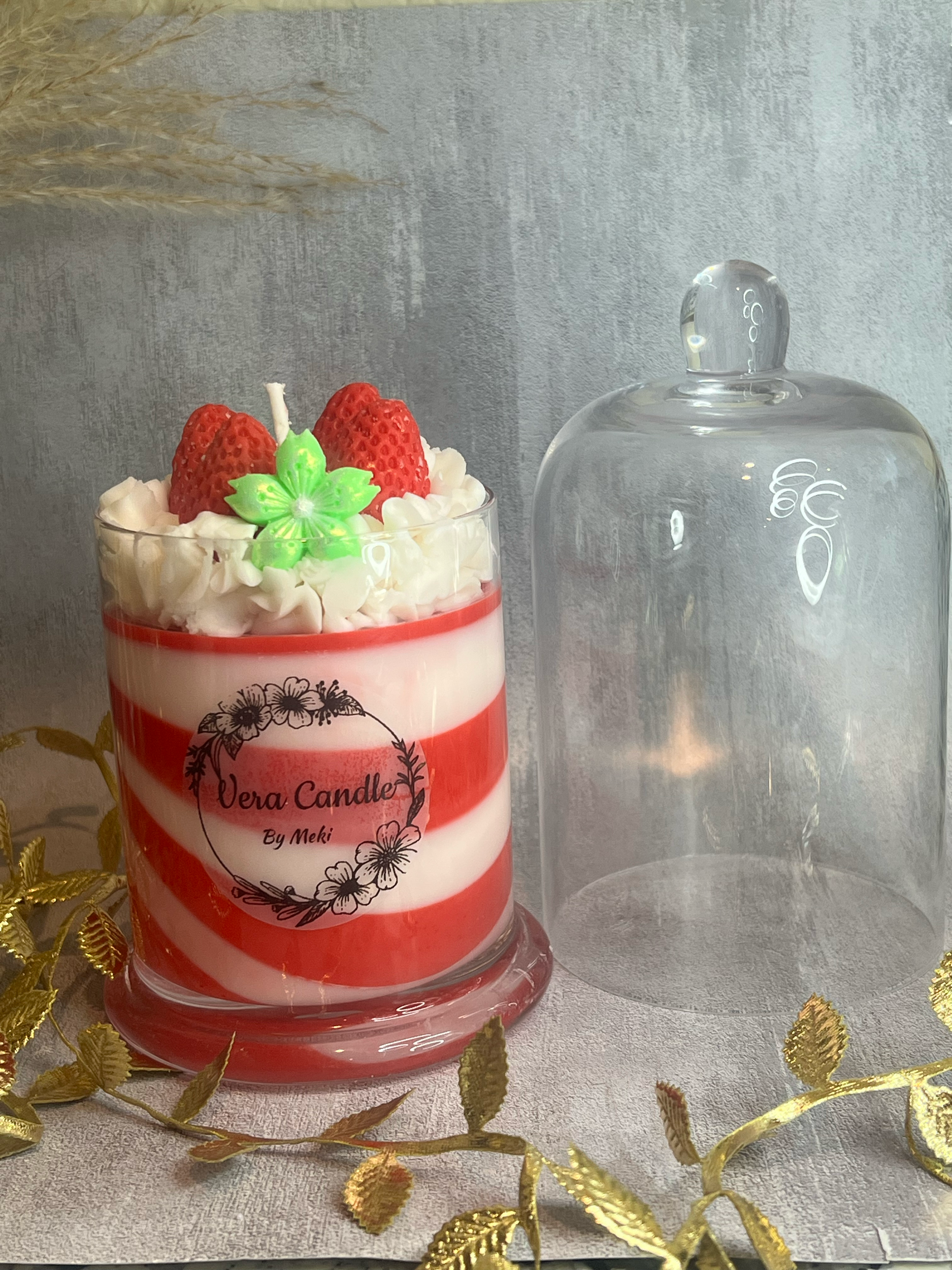 A red and white striped candle with a label that says 'Vera Candle by Meki,' topped with whipped cream, two strawberries, and a green candle shaped like a flower, placed next to an empty glass dome on a surface decorated with gold leaves.
