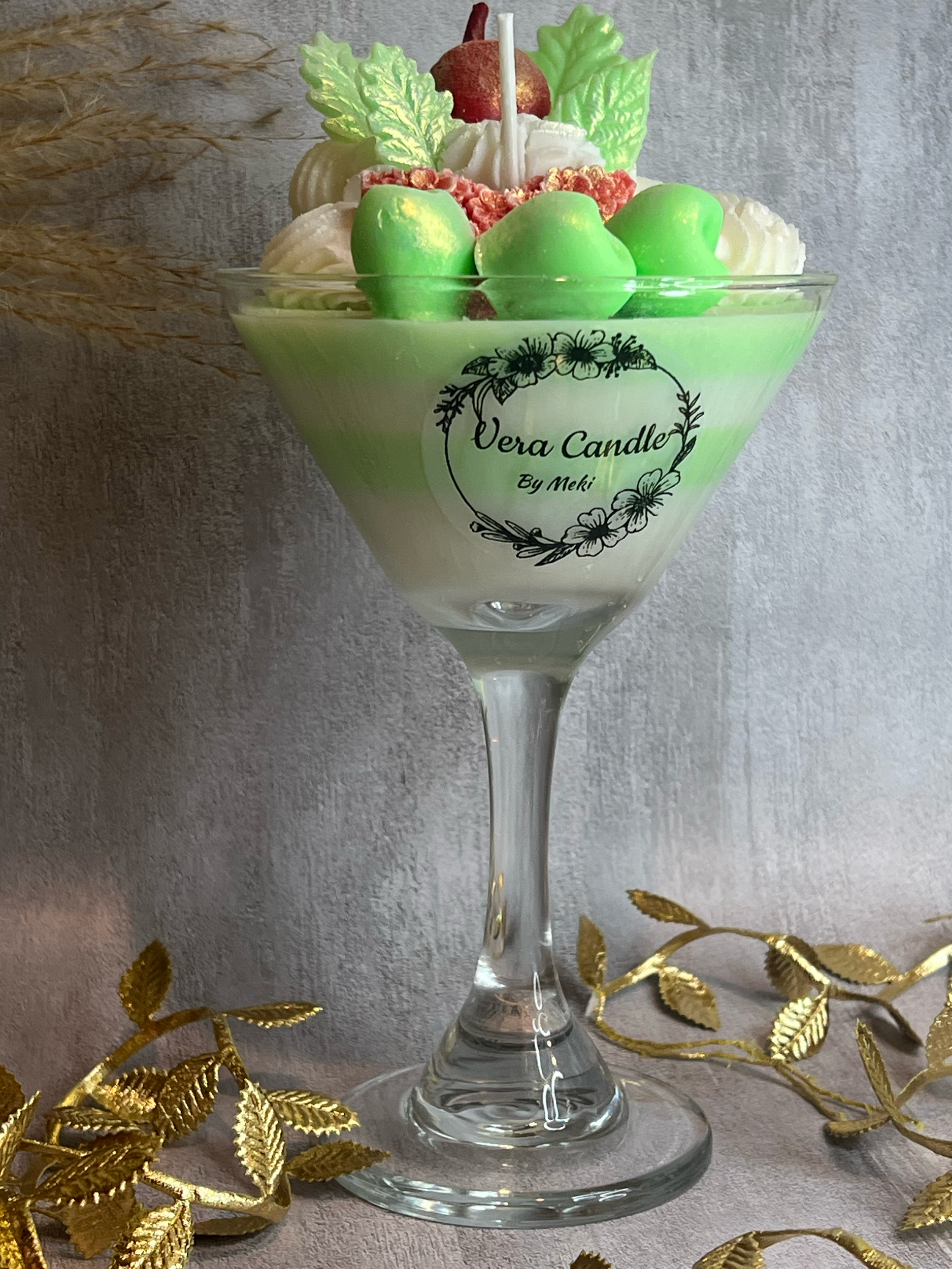 A decorative candle in a martini glass with green, white, and red colors, topped with green chocolates, a red apple, and light green leaves, with a label that reads "Vera Candle By Meki". Gold decorative leaves are placed around the glass.