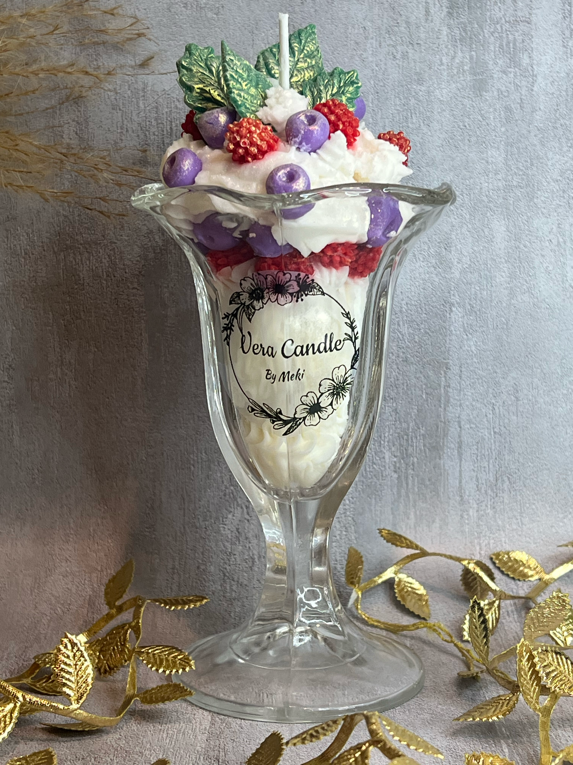 A decorated glass candle holder with a white candle, filled with miniature faux berries and leaves, featuring a label that says 'Vera Candle by Meki'. The setup includes gold decorative leaves and a textured backdrop.