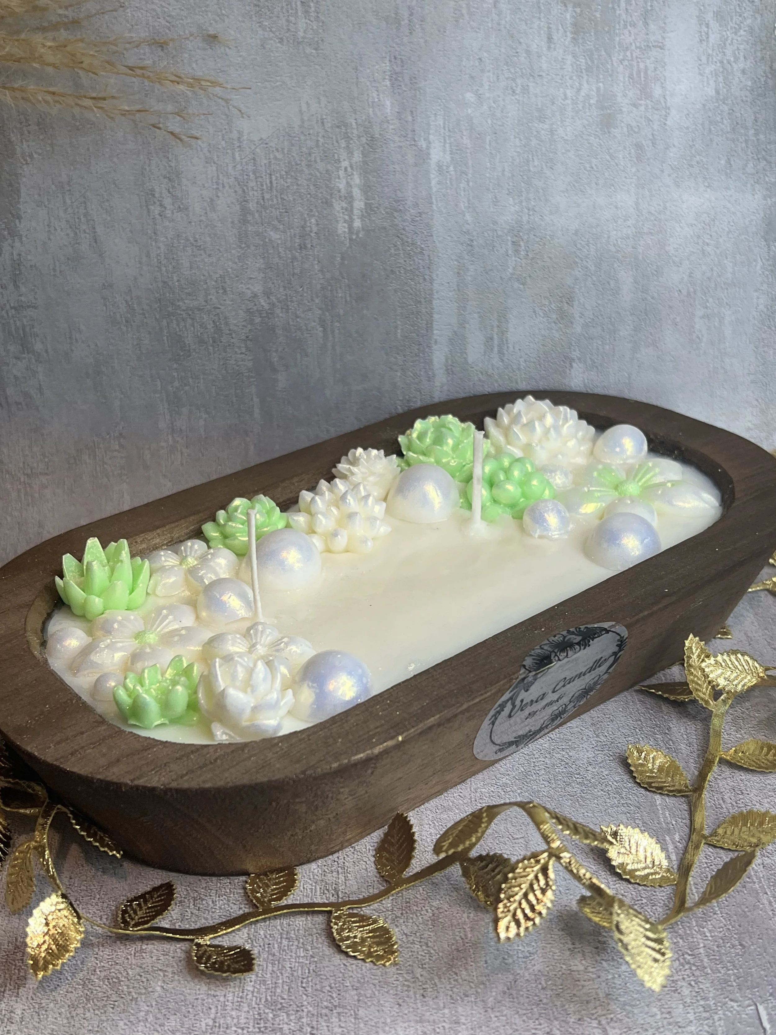 Decorative white and green candles shaped like flowers and succulents arranged on a wooden tray with candles, placed on a beige fabric surface with gold decorative leaves and a silver background.