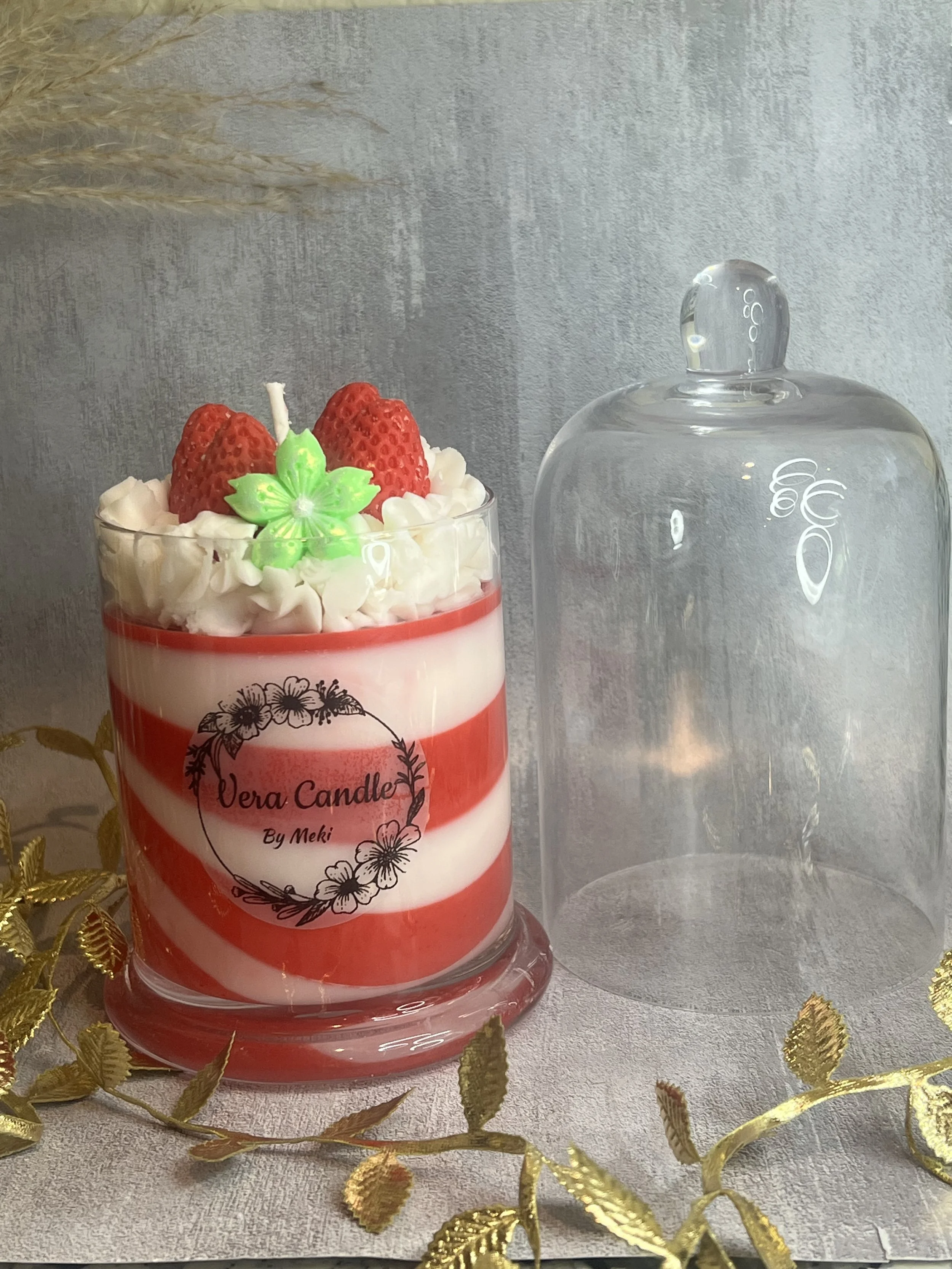 A layered red and white striped candle labeled 'Vera Candle By Meki' with strawberries, whipped cream, and a green decorative flower on top, placed beside an empty glass cover on a table decorated with gold leaves.