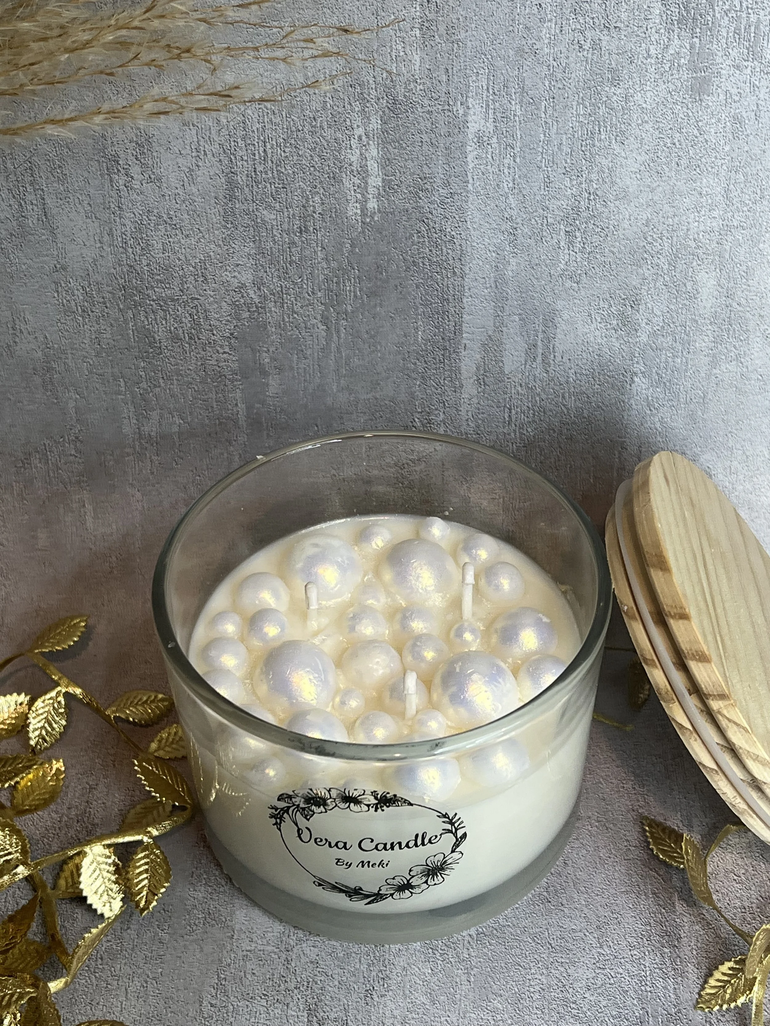 A white scented candle in a glass jar with pearl-like decorations inside, labeled 'Dora Candle by Meki', placed on a textured surface with gold decorative leaves and a wooden lid nearby.