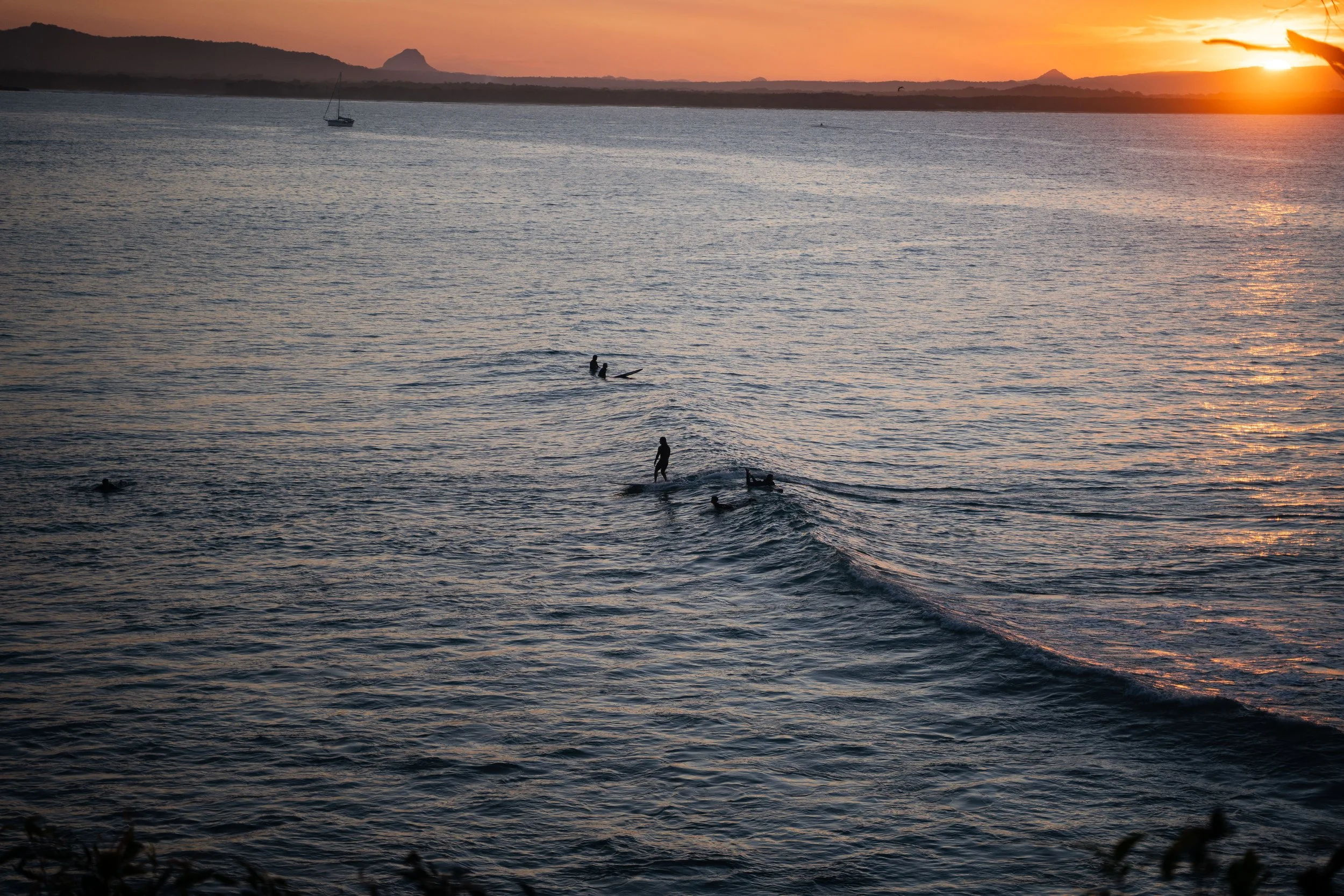 People surfing and swimming in the ocean at sunset with mountains in the background.