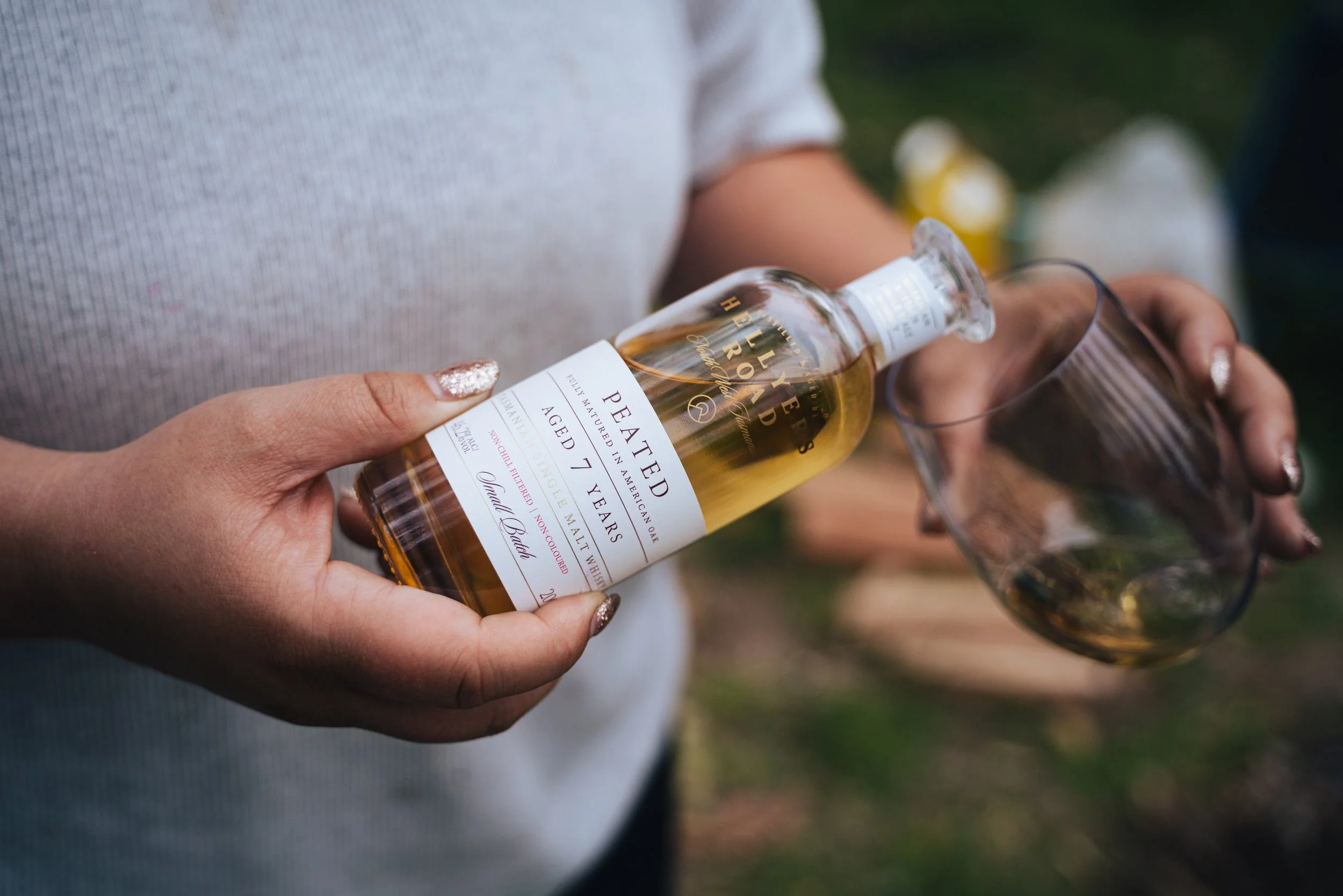 Person pouring whiskey from a bottle into a glass during an outdoor gathering.