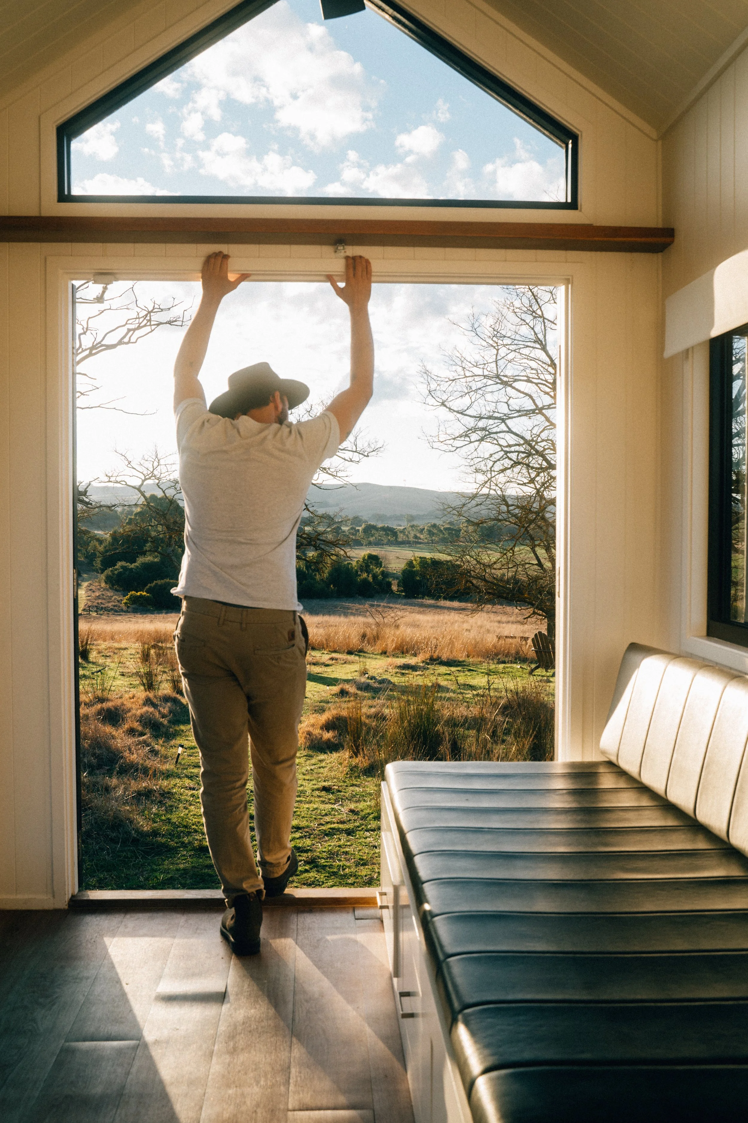 Person opening sliding glass door to outdoor landscape with trees, grassy field, and hills under partly cloudy sky.