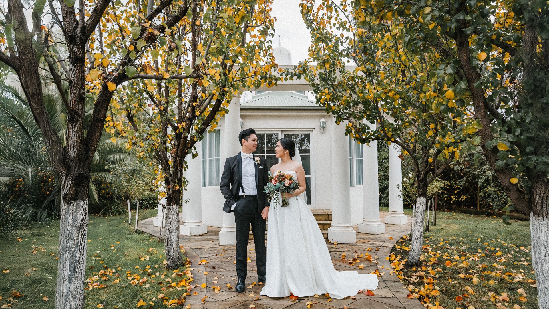 A bride and groom at Sheldon Receptions, standing face to face in a garden with trees and fall foliage, holding hands. The bride is in a white gown holding a large bouquet, and the groom is in a black tuxedo with a boutonniere