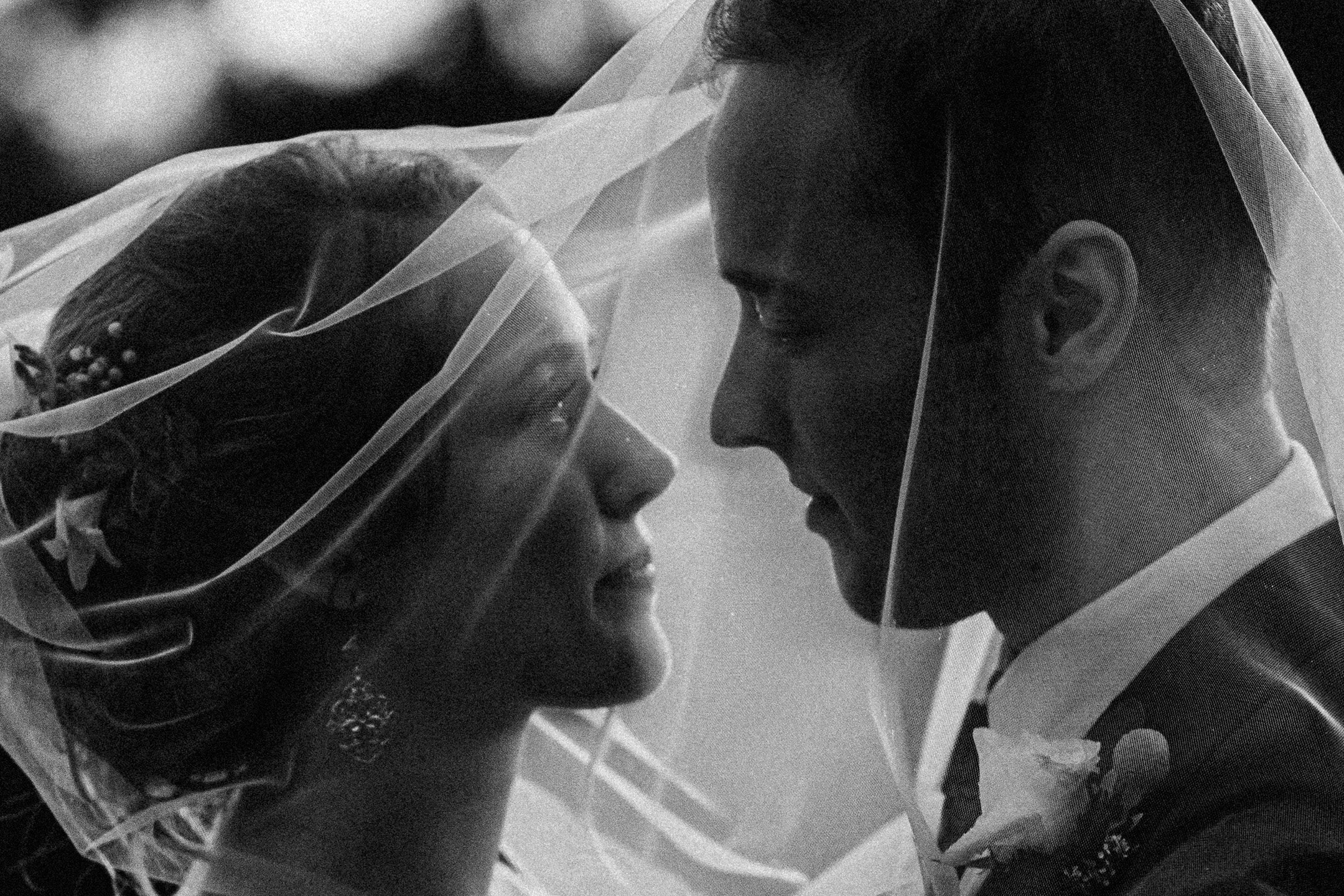 Black and white closeup photo of a bride and groom, face to face, under a veil and smiling at each other.