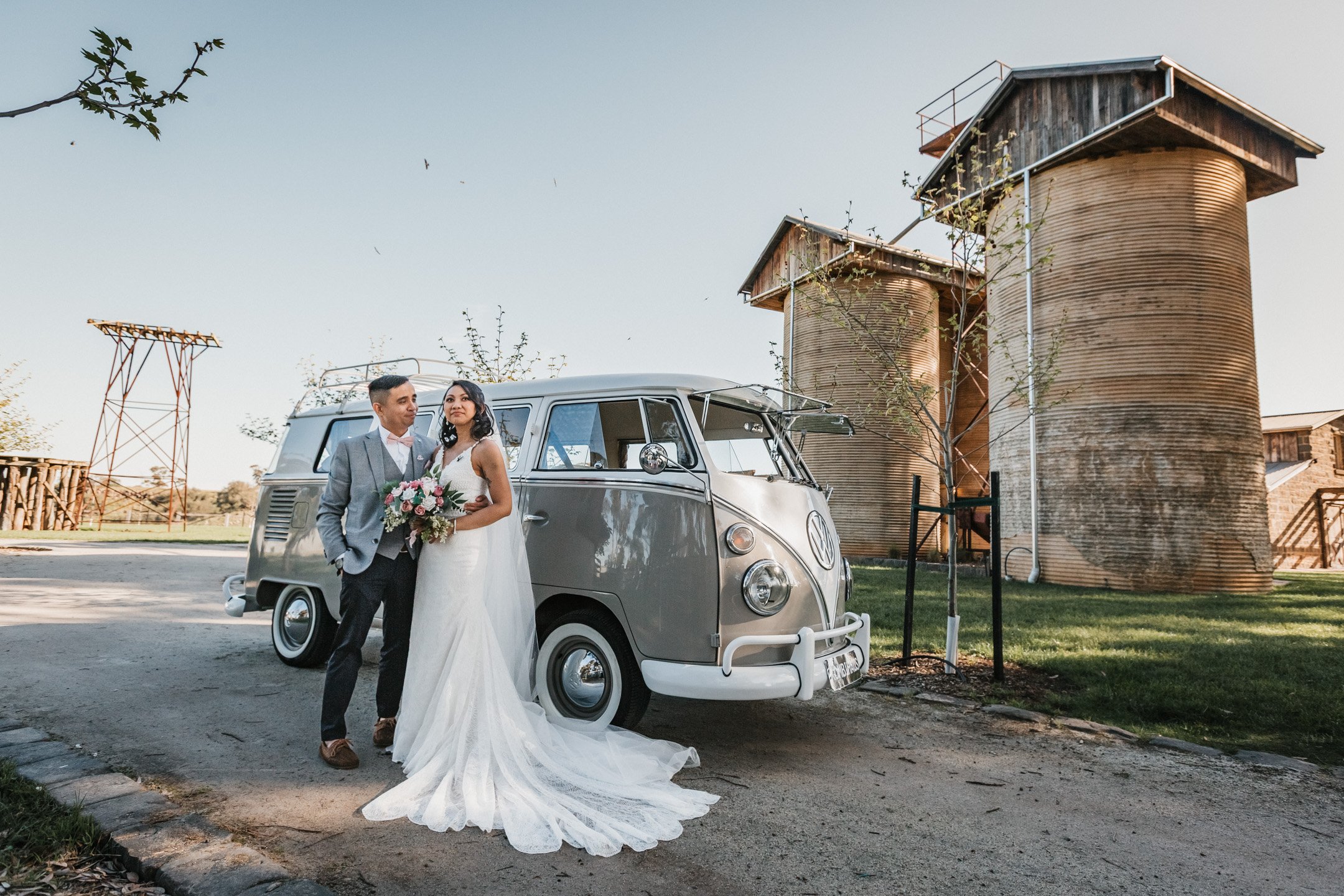 A bride and groom at Warrawong Estate, standing together in front of a vintage Volkswagen camper van, with large silos and a clear sky in the background.