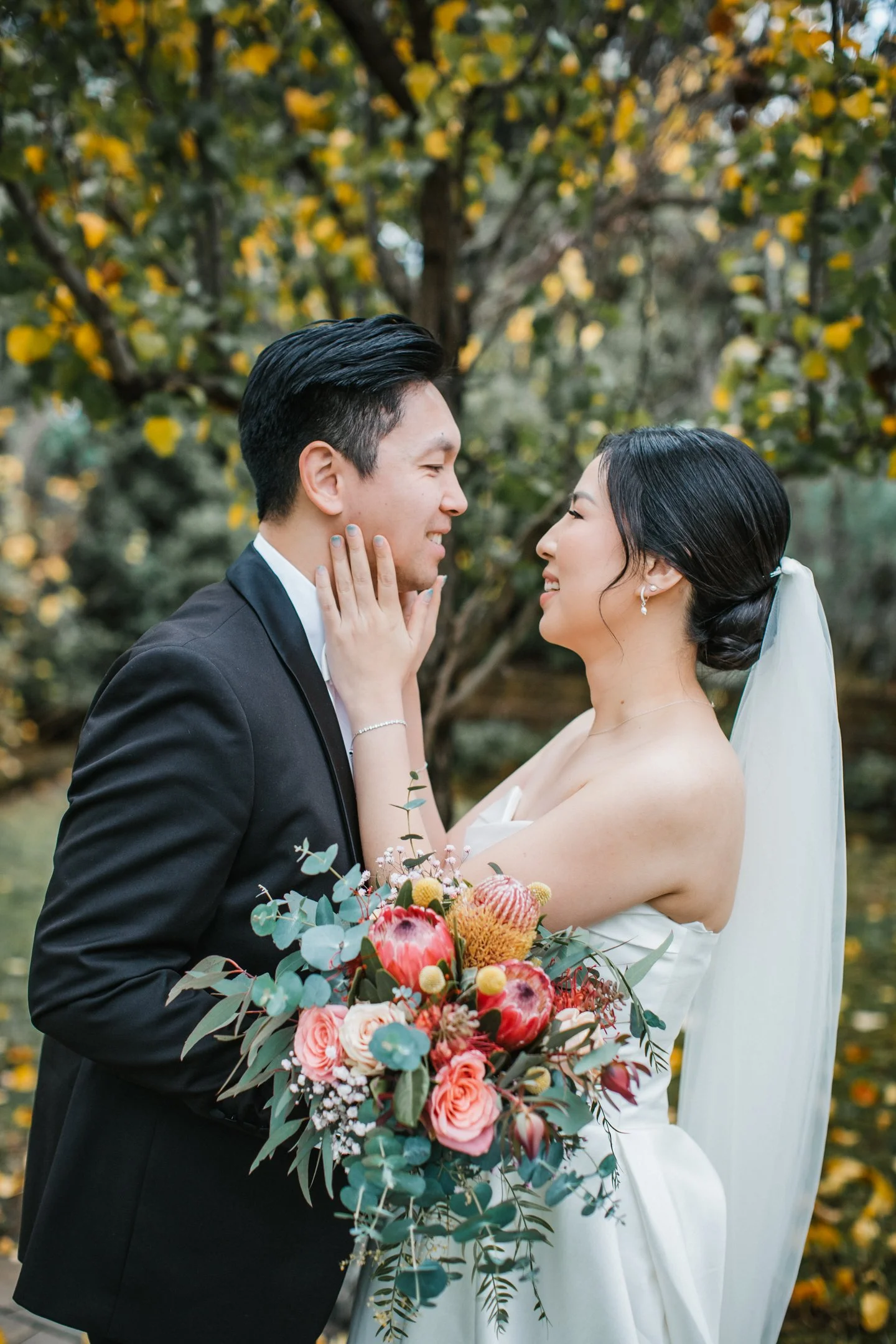 A bride and groom are outdoors, gazing at each other with smiles. The bride holds a bouquet of pink, red, and green flowers, and the groom wears a black tuxedo. The background features yellow and green foliage.