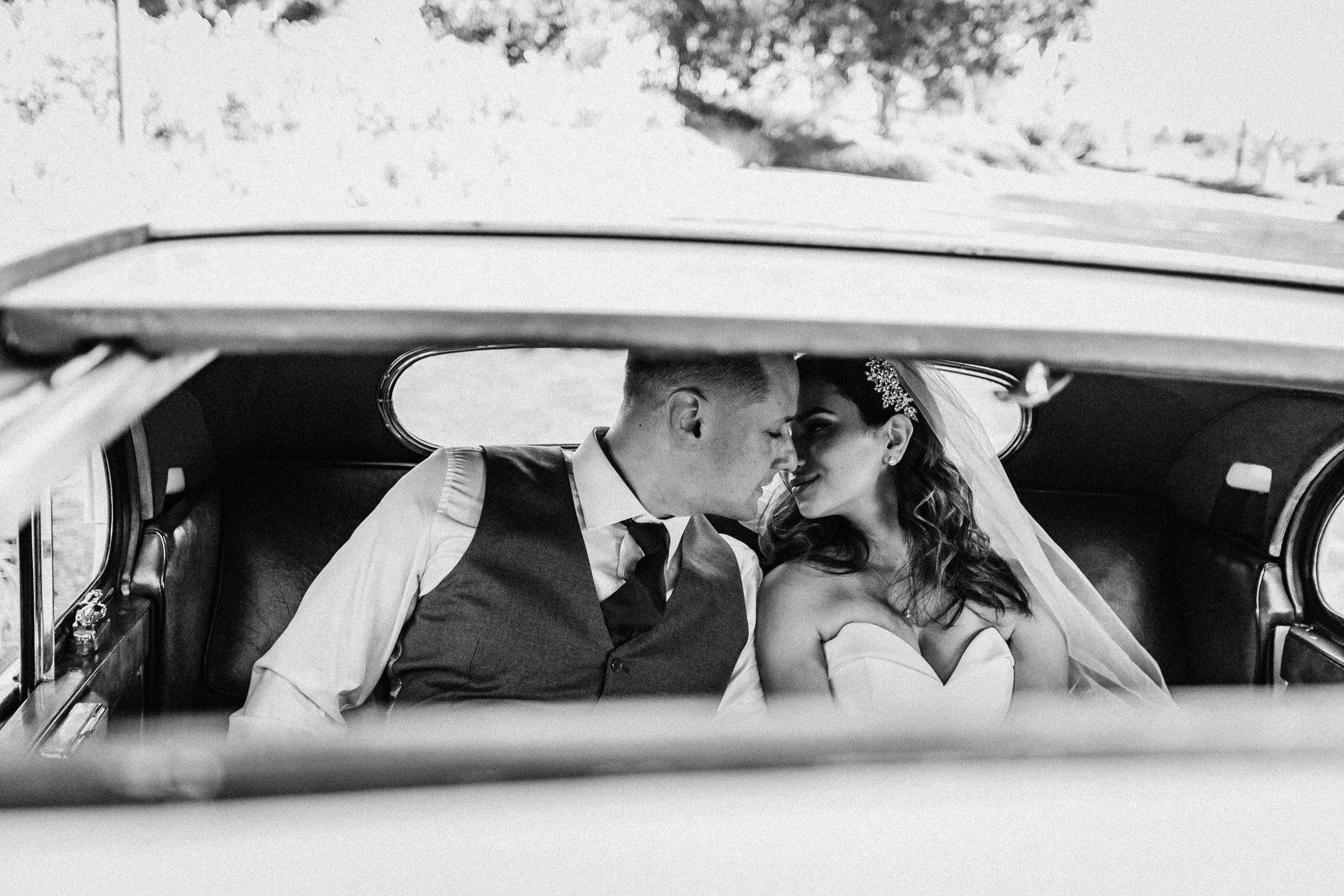 A bride and groom sitting close together in the backseat of a vintage car, with their foreheads touching and eyes closed, sharing an intimate moment.