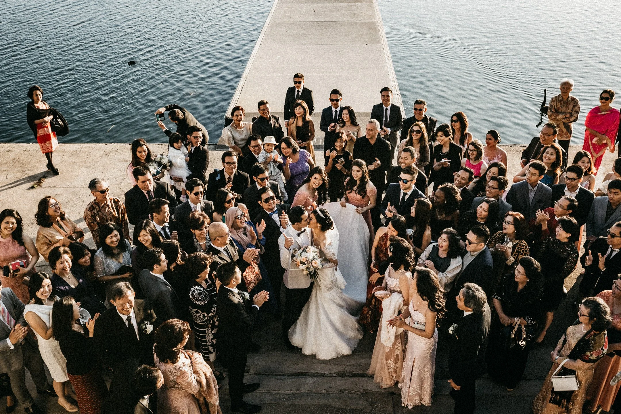 A wedding celebration with the bride and groom kissing among a crowd of guests near a body of water with a pier extending into the water.