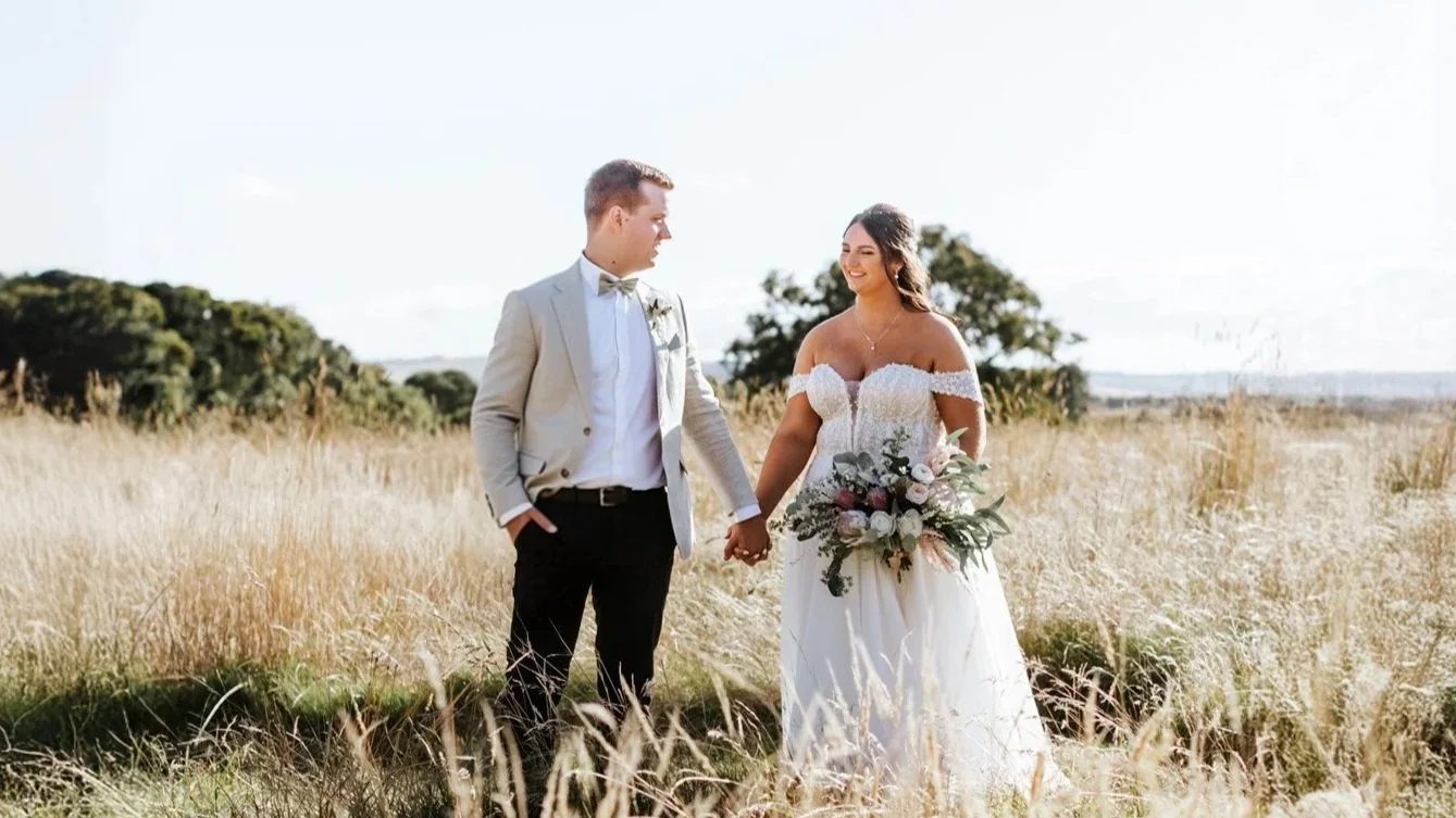 A bride and groom holding hands in a field with tall grass, the bride in a white off-shoulder wedding gown holding a bouquet, and the groom in a light-colored suit with a bow tie, during their outdoor wedding.