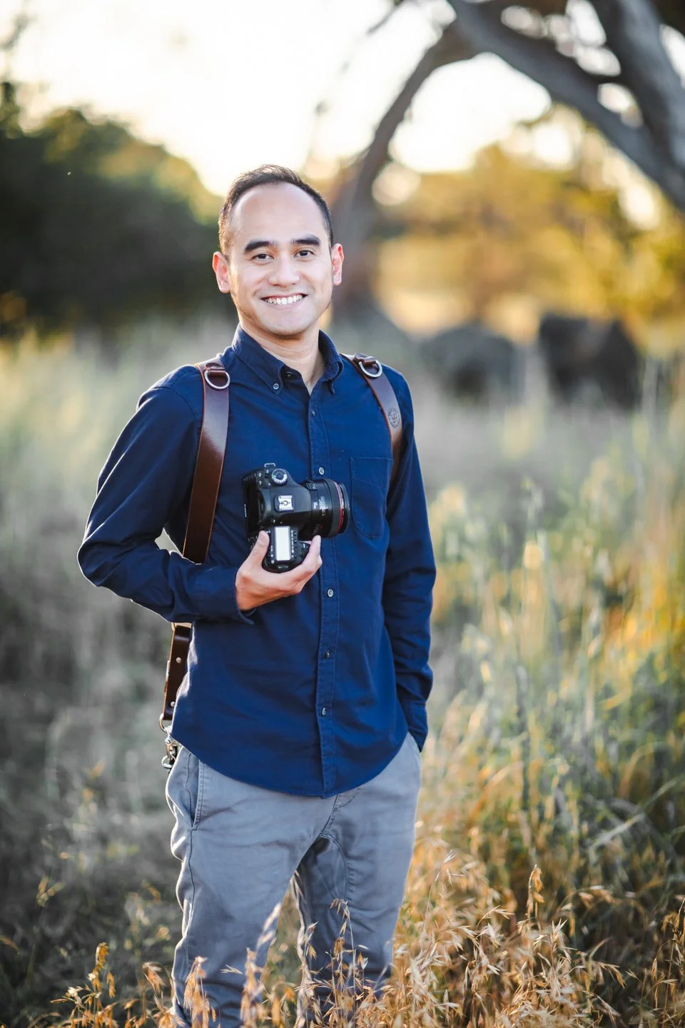 Widi, a photographer, in a dark blue shirt and light gray pants standing outdoors in a field, holding a camera, with a backpack, set against a background of trees and golden sunlight.
