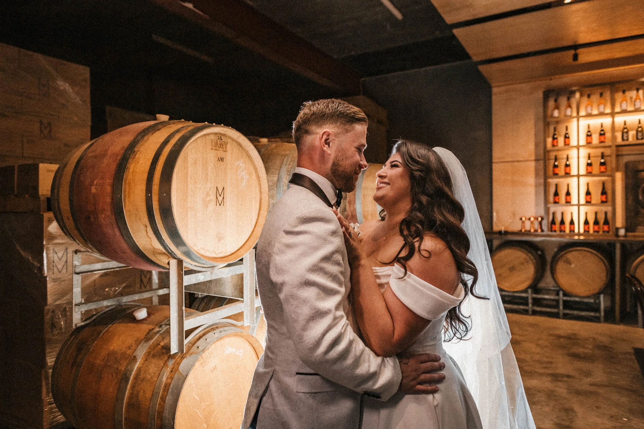 A bride and groom share a romantic moment in a rustic whiskey bar, surrounded by barrels and shelves of whiskey bottles.