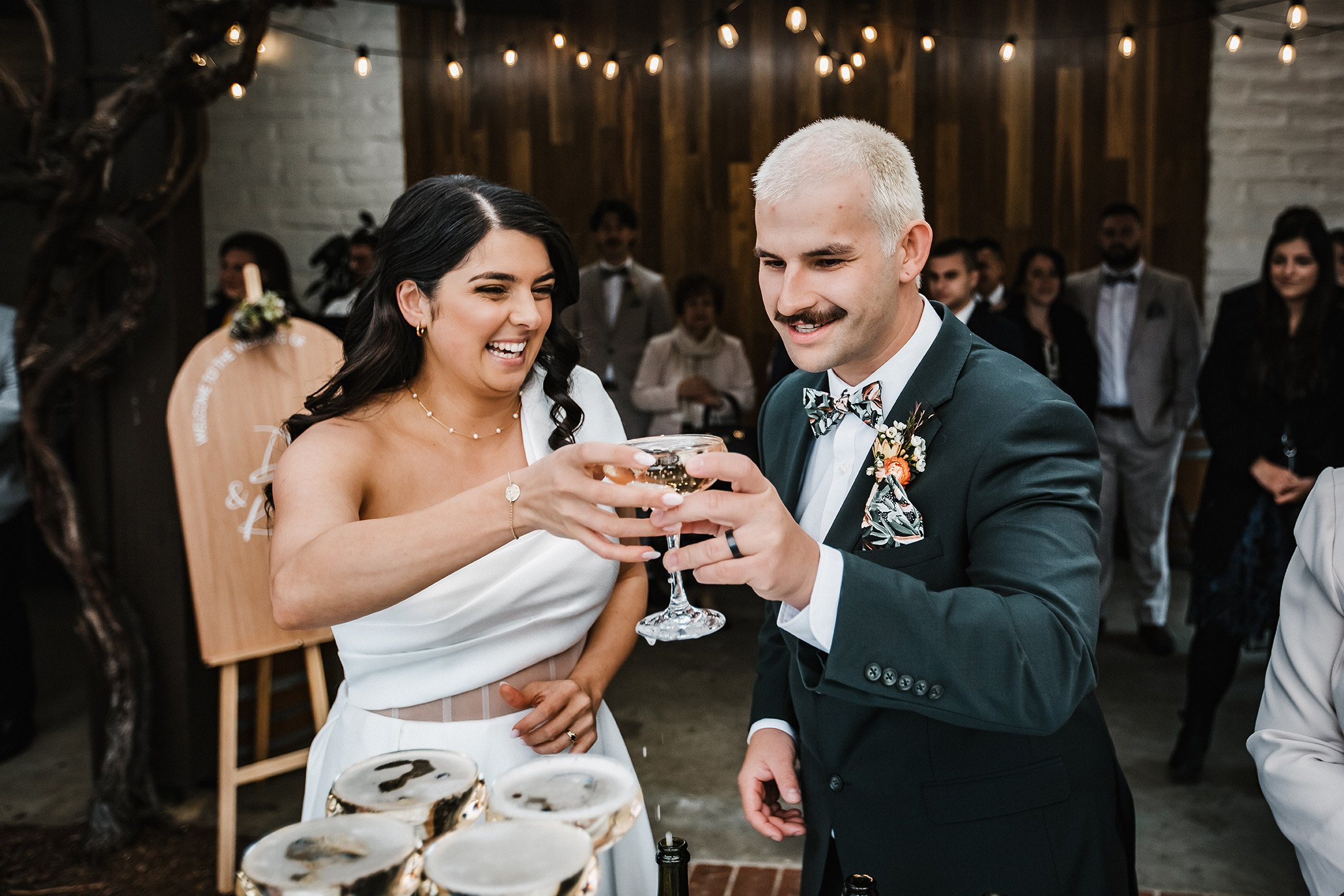 A smiling bride and groom at a wedding celebration toasting with a glass of drink, surrounded by wedding guests in a decorated indoor venue.