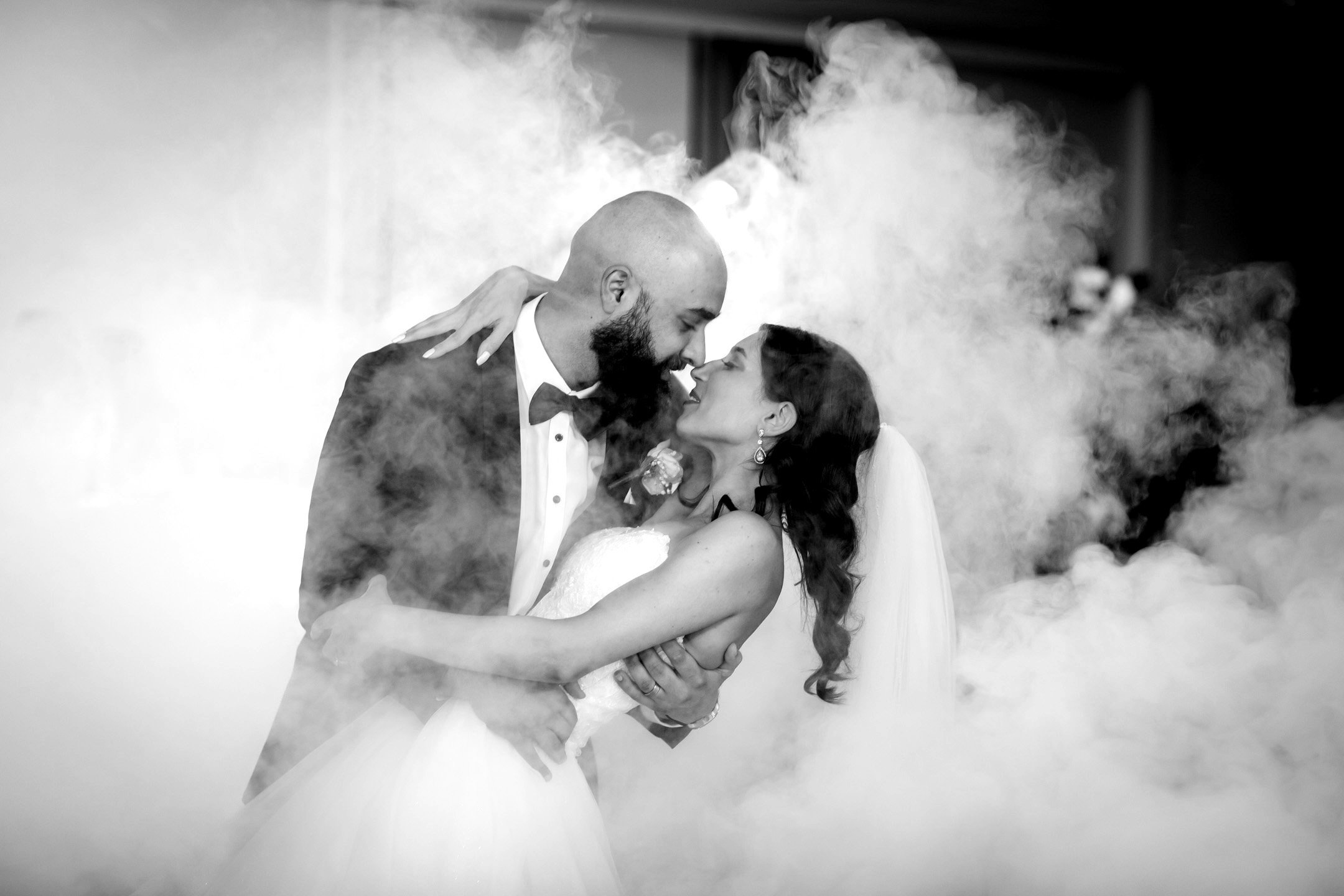A black and white photo of a newlywed couple dancing, surrounded by fog or smoke, with the groom in a tuxedo and the bride in a strapless wedding dress, leaning into each other with closed eyes.