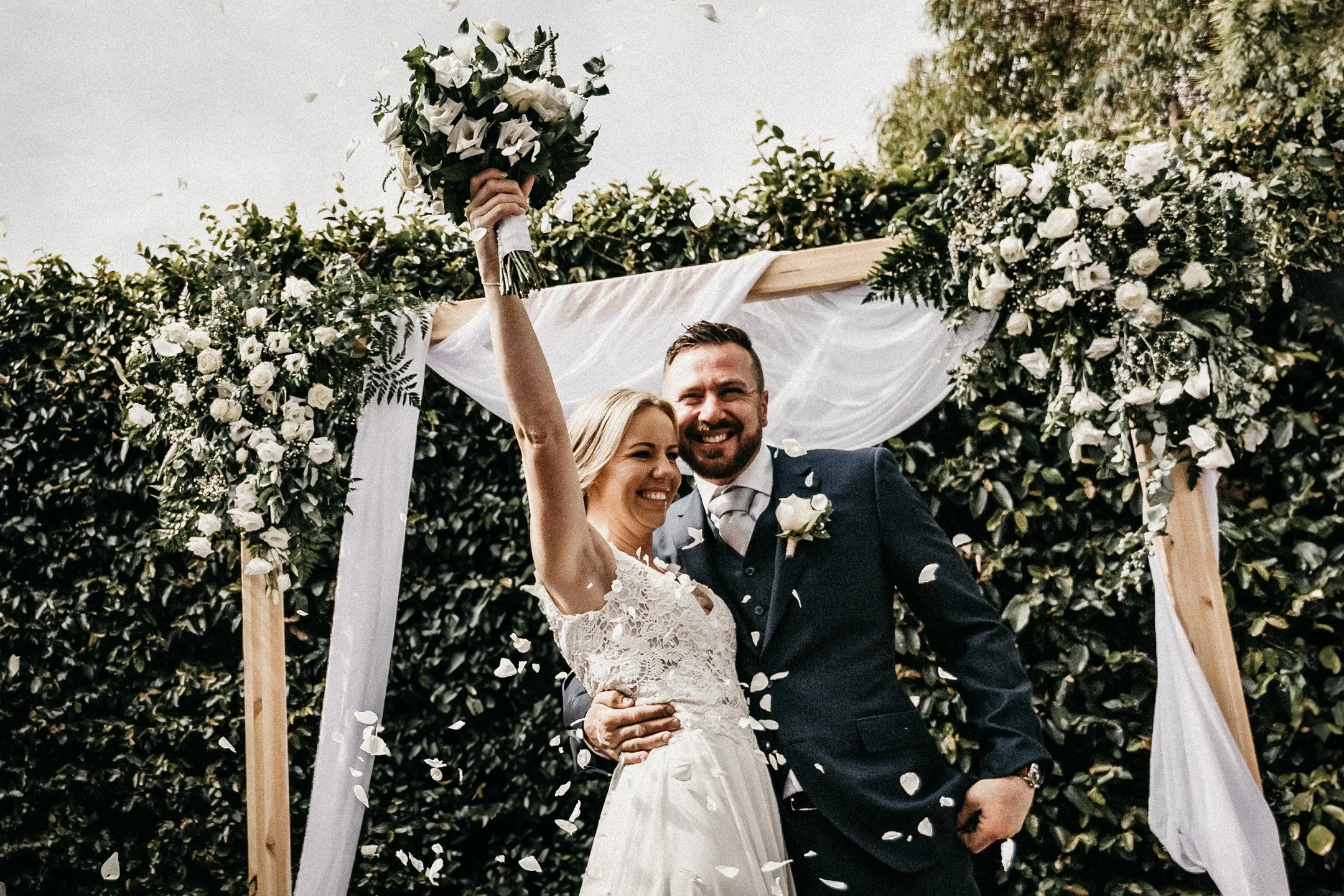 A happy bride and groom celebrating their wedding outdoors, with the bride holding a bouquet of flowers in the air. They are smiling and embracing under a decorated wooden arch with greenery and white flowers. Confetti is falling around them.