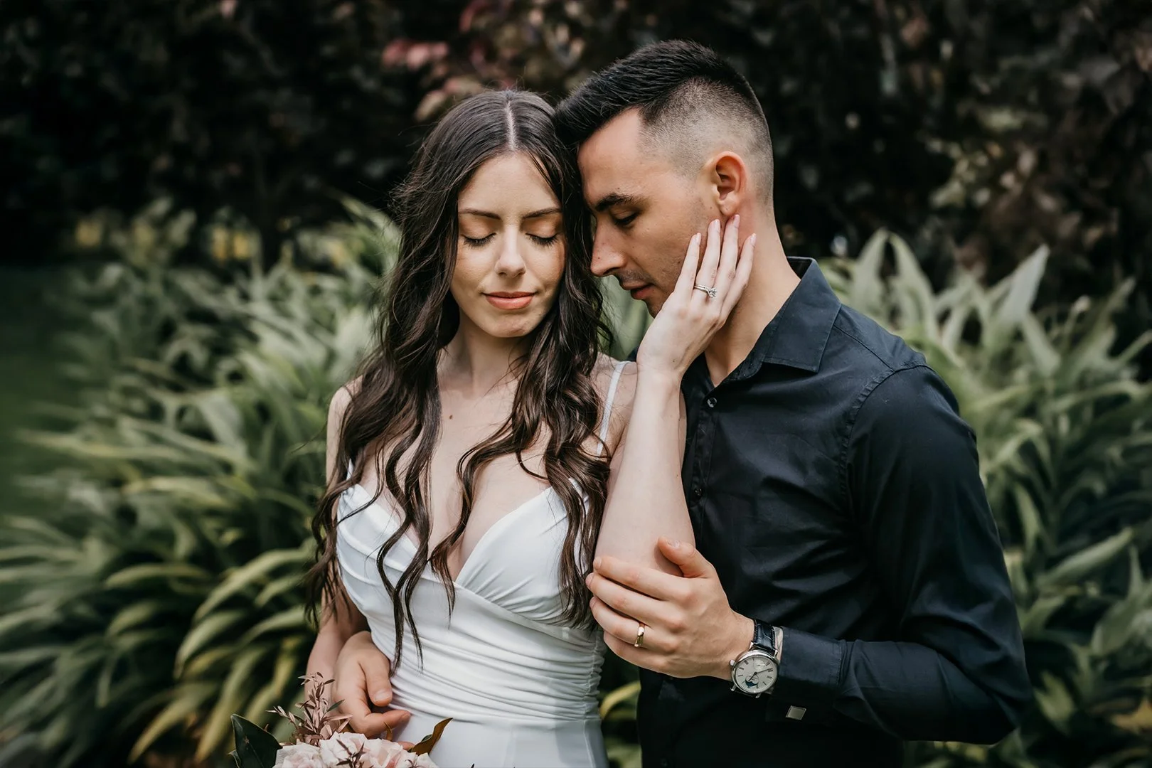 A young couple stands close together outdoors, eyes closed, with the woman's hand gently resting on the man's face, wearing wedding rings; she is in a white dress holding a bouquet, he is in a dark shirt, surrounded by lush greenery.