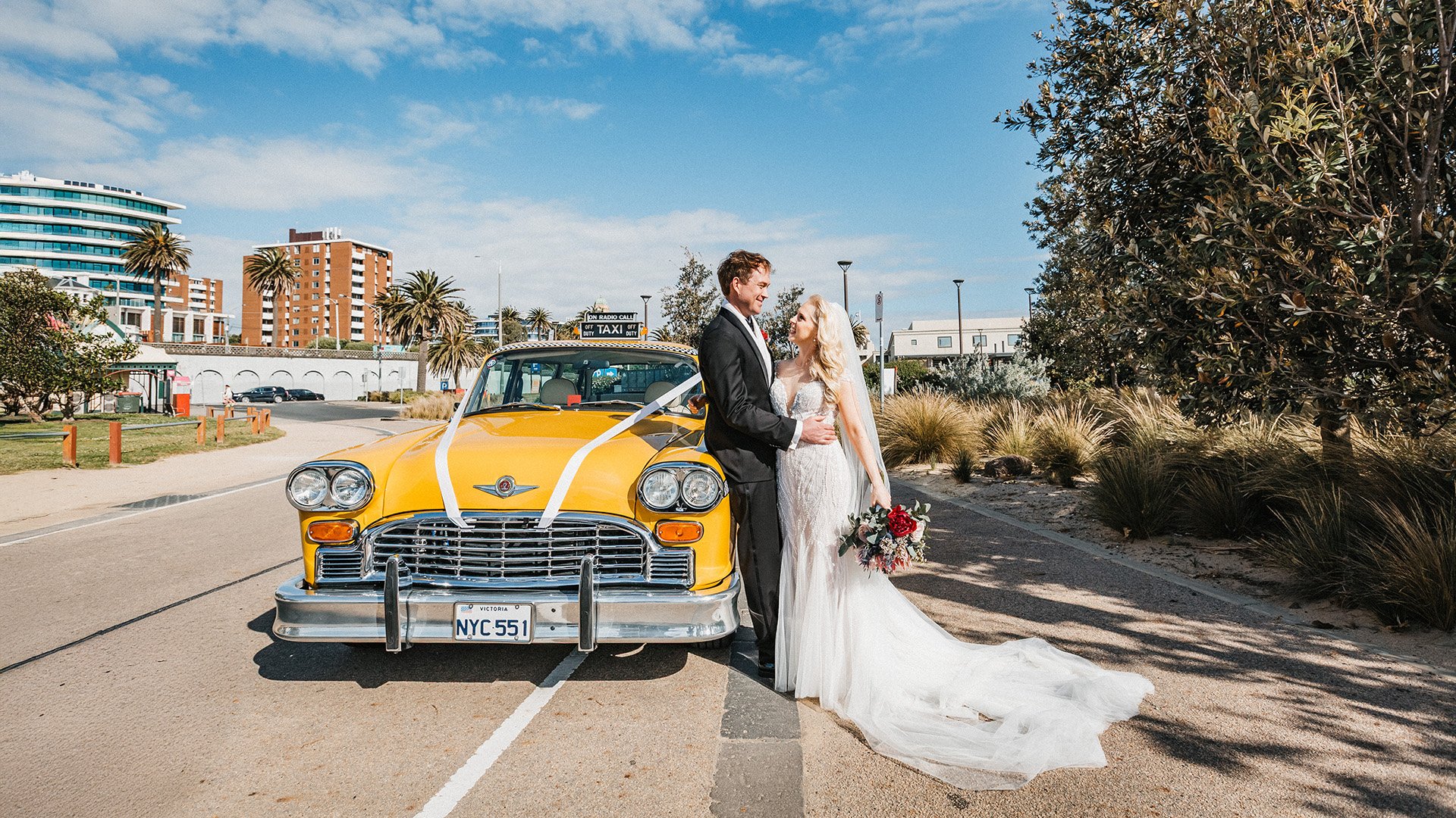 Wedding couple in wedding dress and tuxedo standing next to a yellow taxi decorated with white ribbons, holding each other and smiling outdoors on a city street with modern buildings and palm trees.