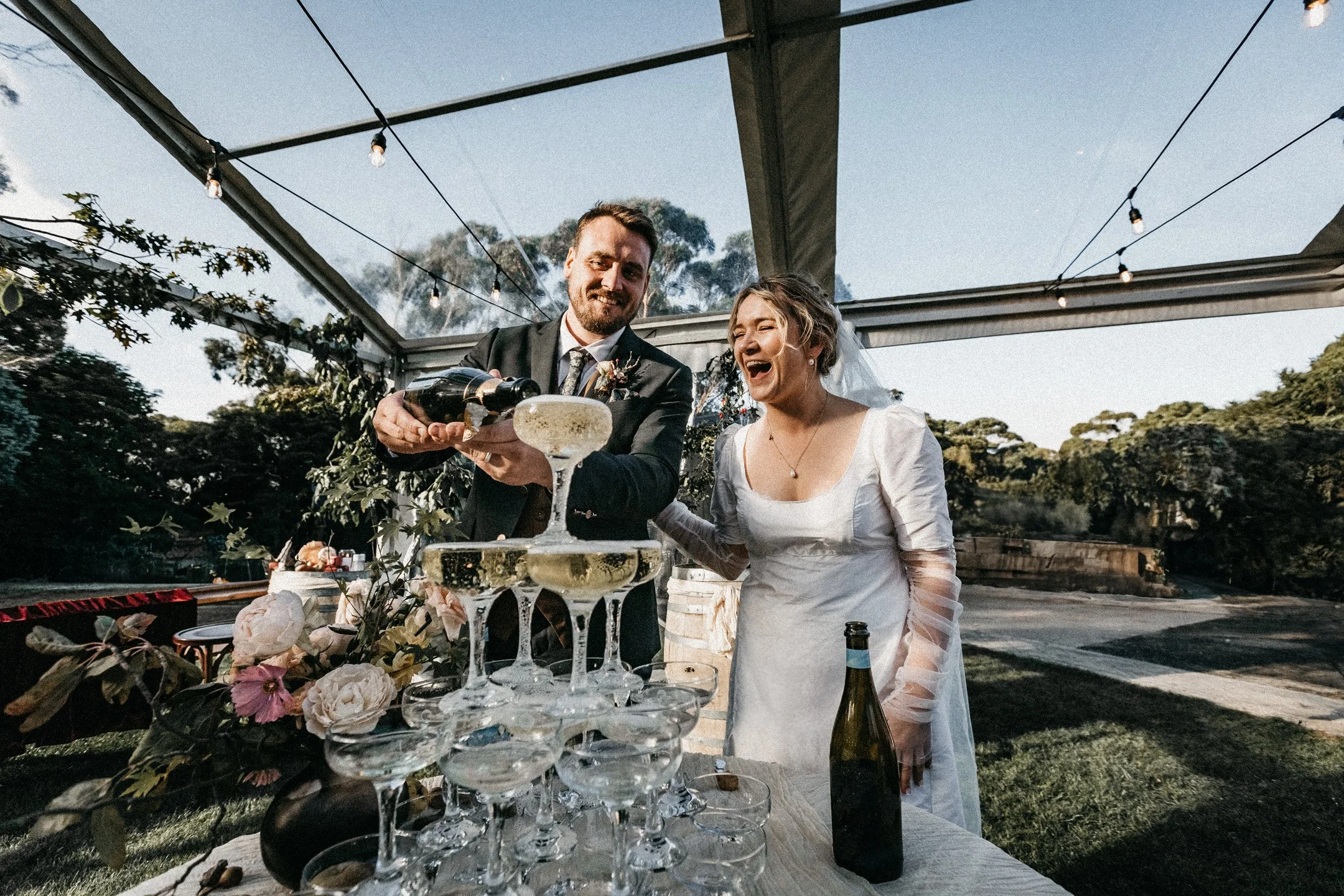 A newlywed couple celebrates by pouring champagne into a tower of glasses outdoors under a canopy, with trees and a clear sky in the background.