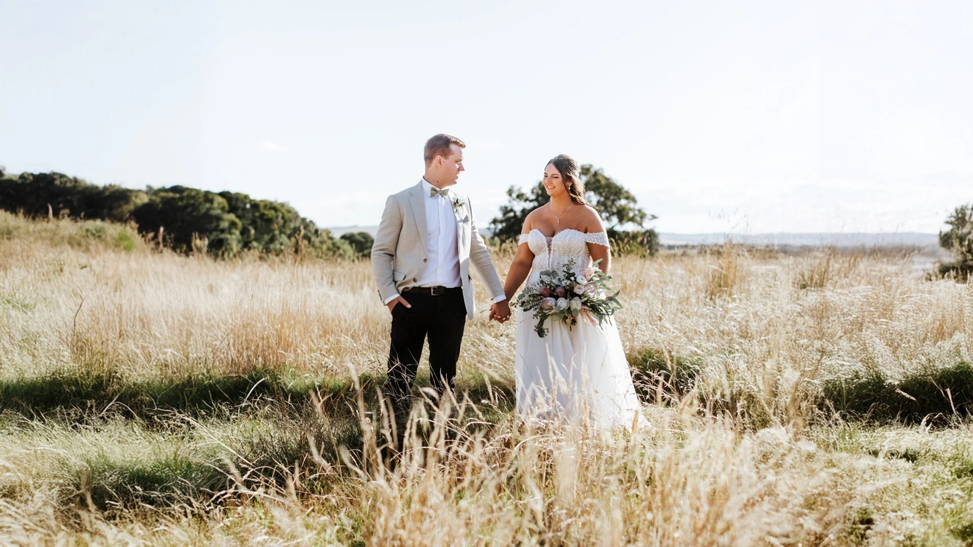 A bride and groom holding hands in a grassy field during a wedding photoshoot, with trees and a clear sky in the background.
