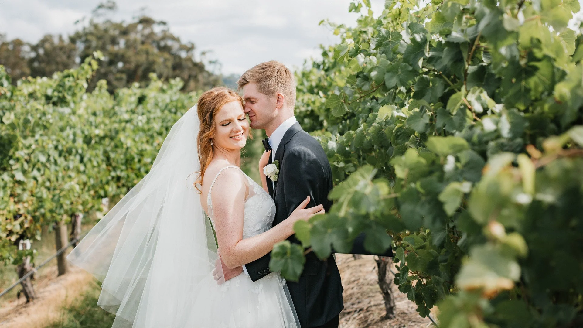 A bride and groom sharing a tender moment in a vineyard at The Vines of Yarra Valley, with the bride smiling and the groom gently leaning in, surrounded by lush green grapevines.