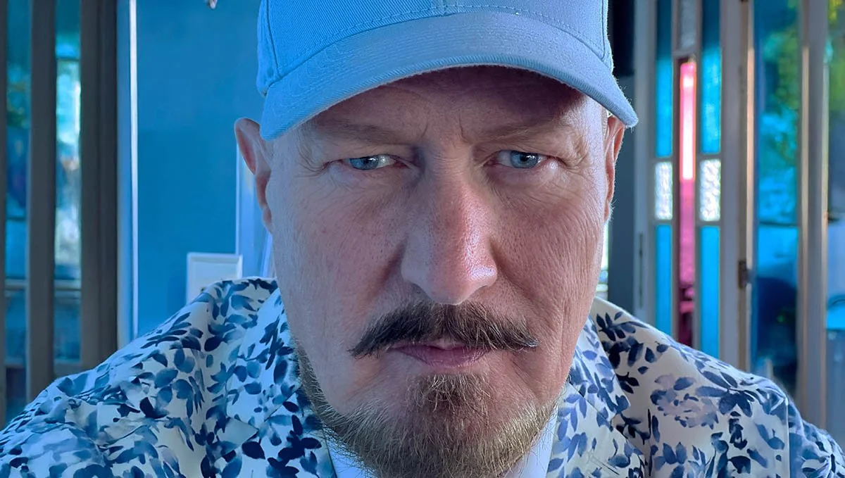 Costume designer Kjell Nordstrøm looks intensely into the camera with a serious expression. He is wearing a light blue cap and a white suit with a pattern of small blue leaves.