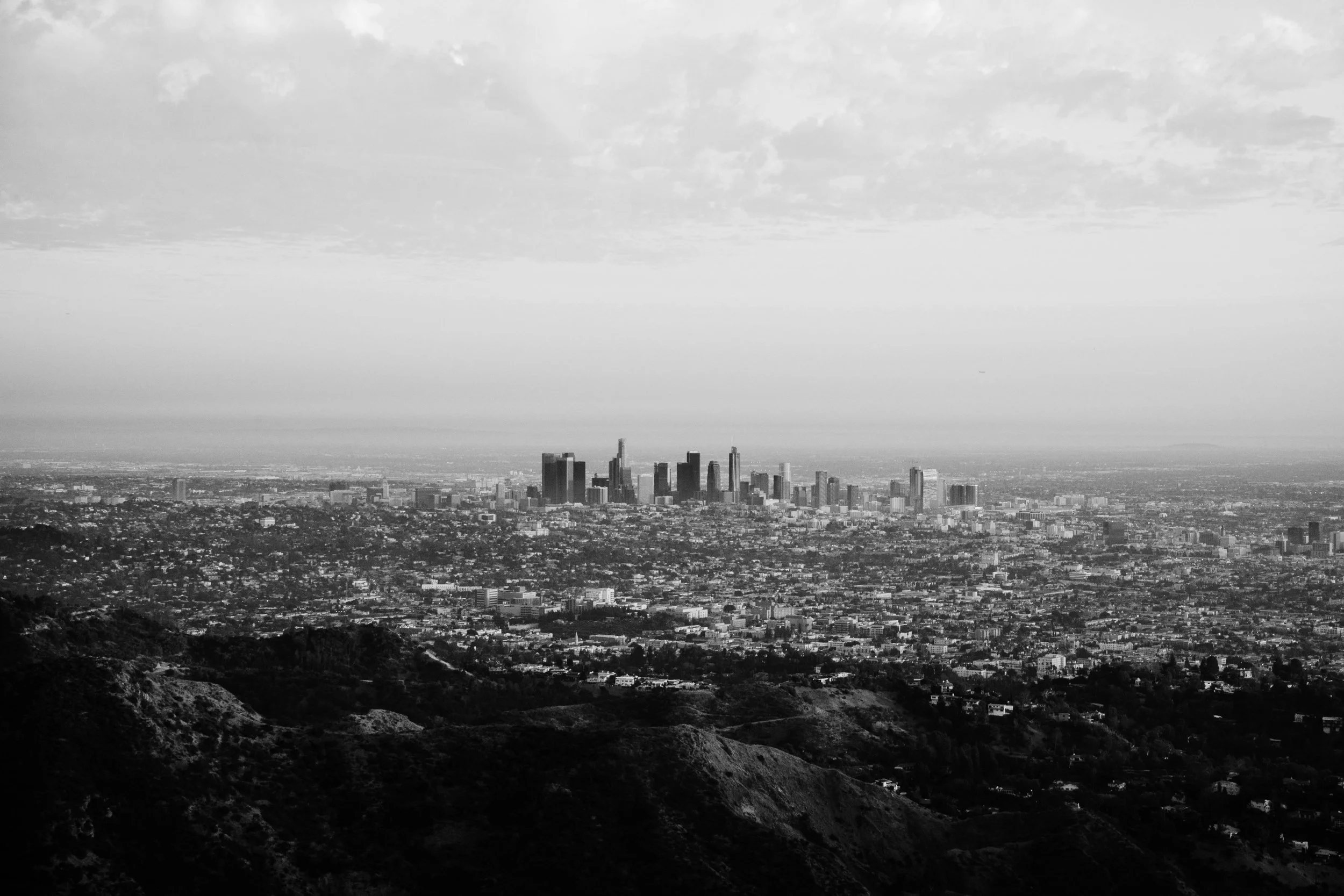Black and white photo of the Los Angeles skyline taken from a distance, showing tall buildings and city spread out under a cloudy sky.