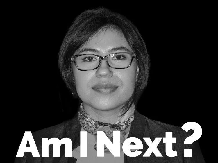Black and white photo of a woman with short hair, glasses, and a patterned scarf, looking at the camera with the text "Am I Next?" in bold white letters at the bottom.
