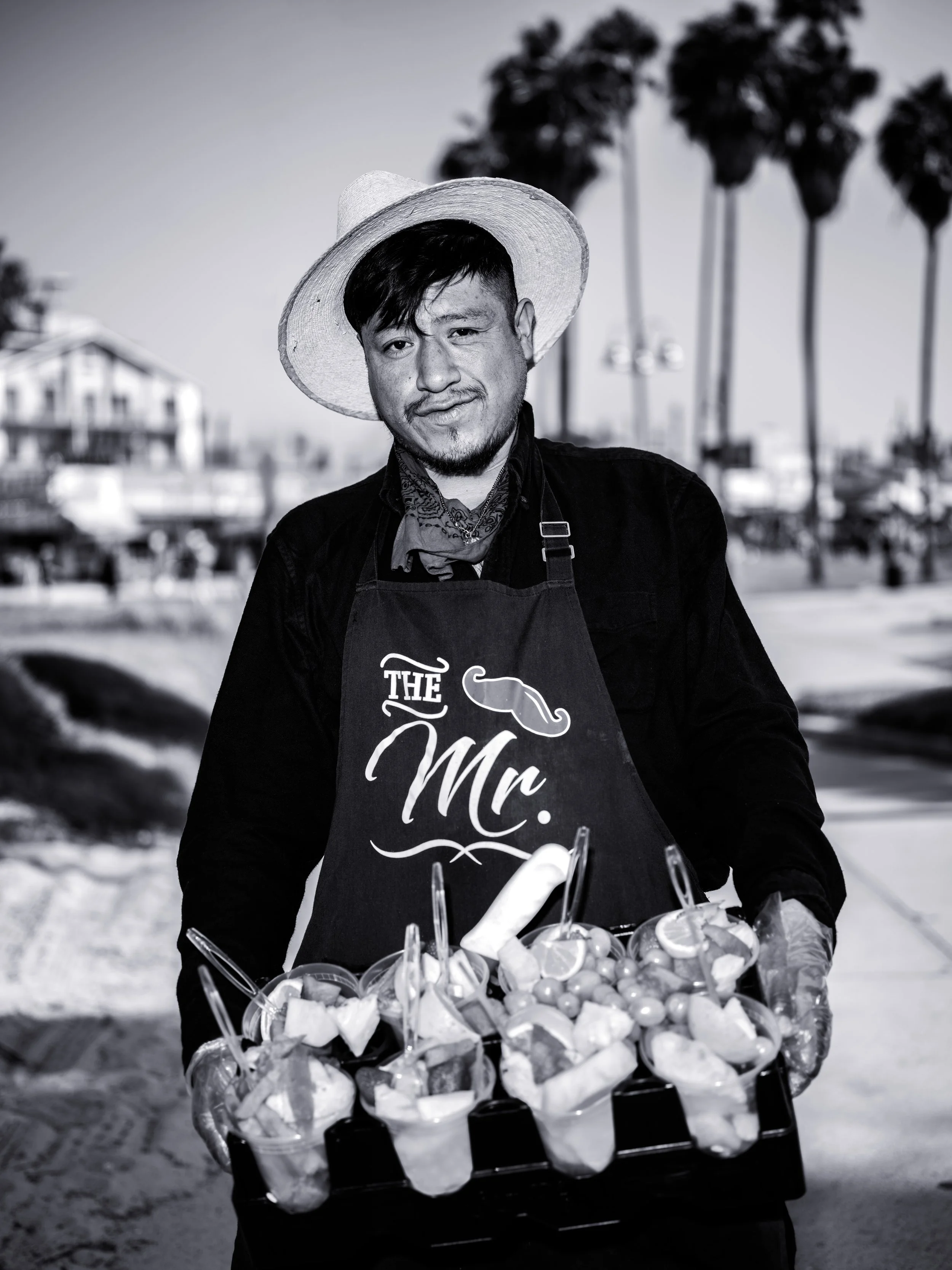 A man wearing a straw hat, black shirt, and apron labeled 'The Mr.' stands outdoors on a beach with palm trees in the background, holding a tray of cups with fruit and utensils.
