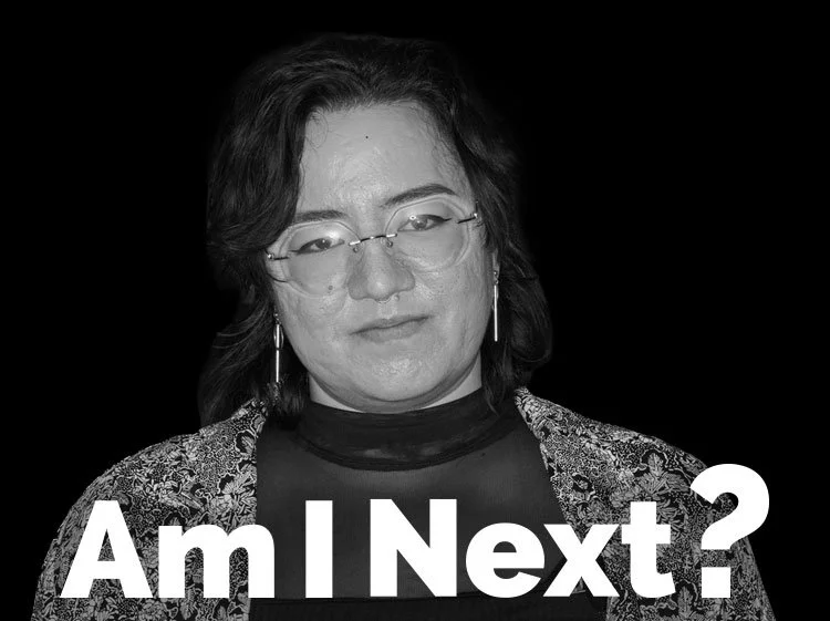A woman with glasses and earrings, wearing a patterned jacket and a black top, looking down with a contemplative expression, captioned 'Am I Next?' in large white letters.