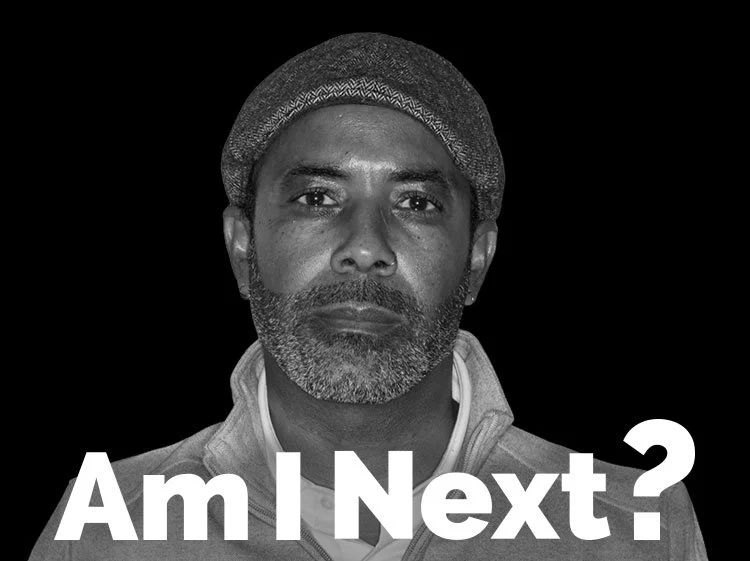 A black-and-white portrait of a middle-aged man with a beard and mustache, wearing a beanie and a zip-up jacket, looking directly at the camera with a serious expression. Text overlay reading "Am I Next?" is at the bottom of the image.