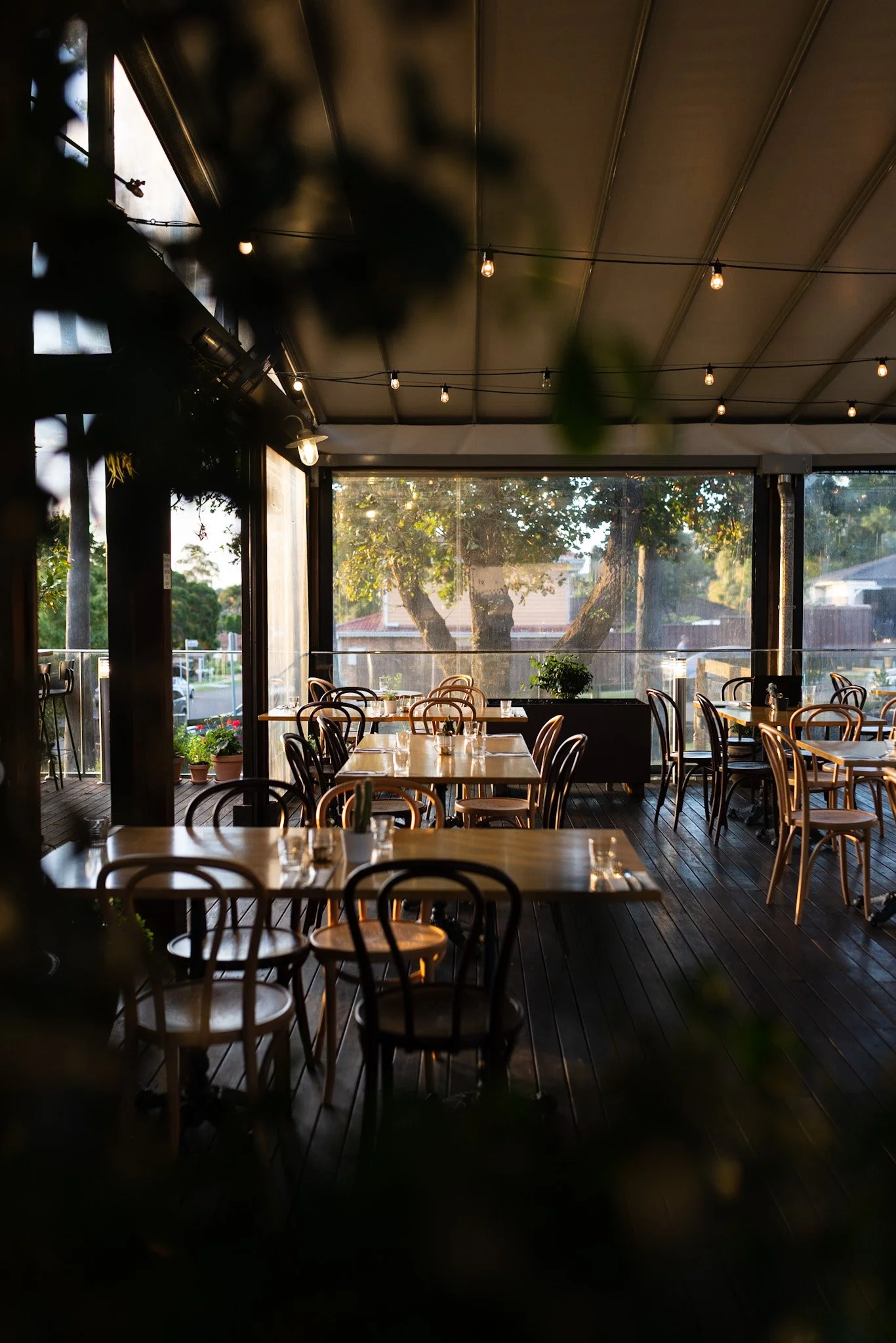 Sunlit empty restaurant patio with wooden tables and chairs, string lights overhead, and greenery outside.