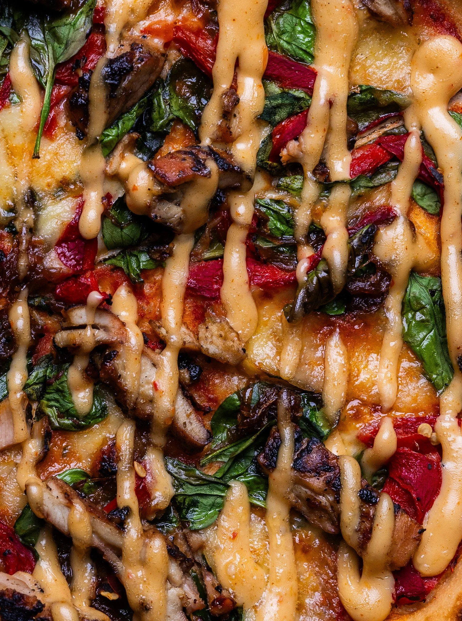 Close-up of a pizza with melted cheese, grilled vegetables, spinach, red bell peppers, and drizzled sauce.