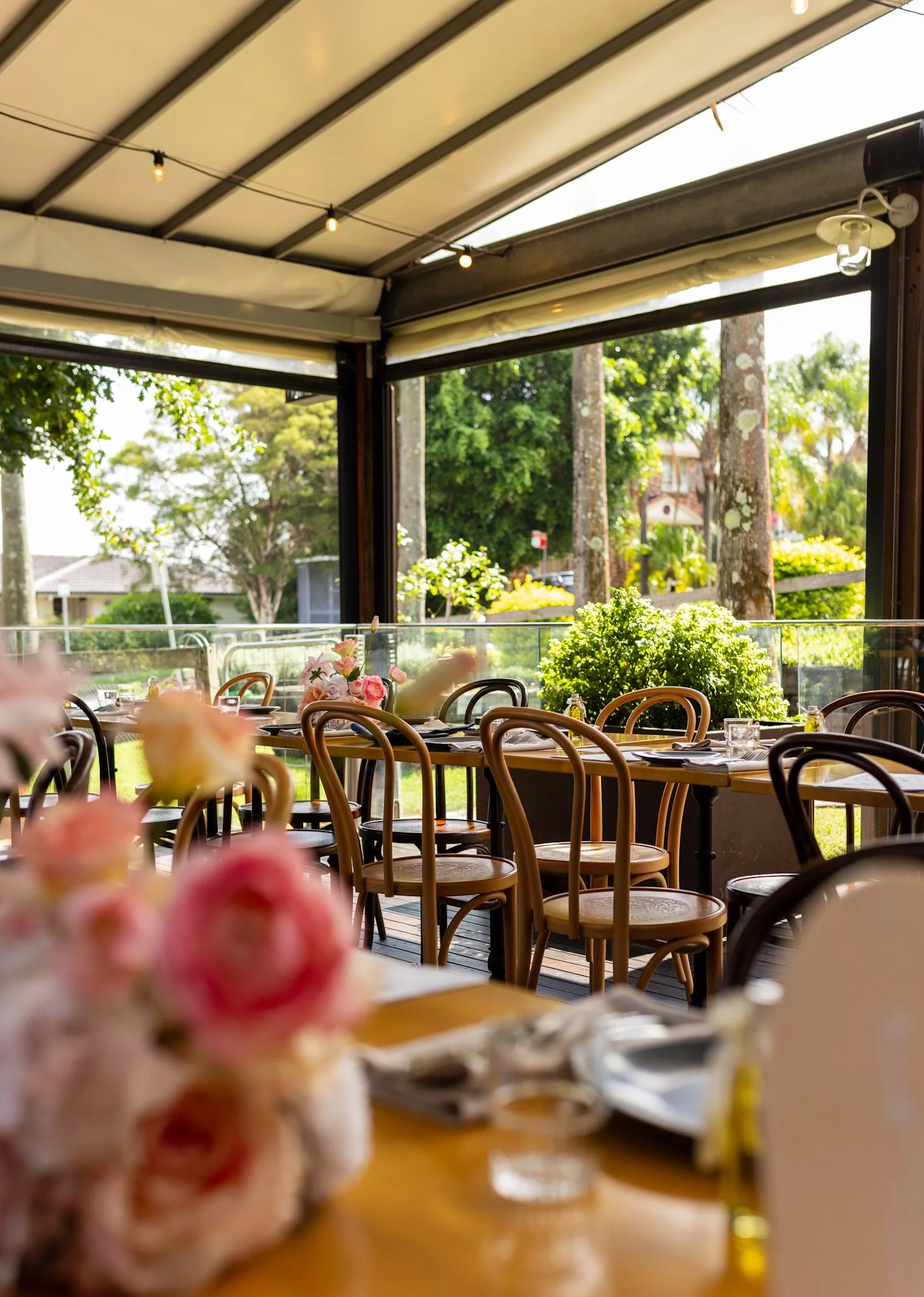 Indoor dining area overlooking a lush green garden with trees, set for a meal with plates, glassware, napkins, and decorated with pink and white flowers.