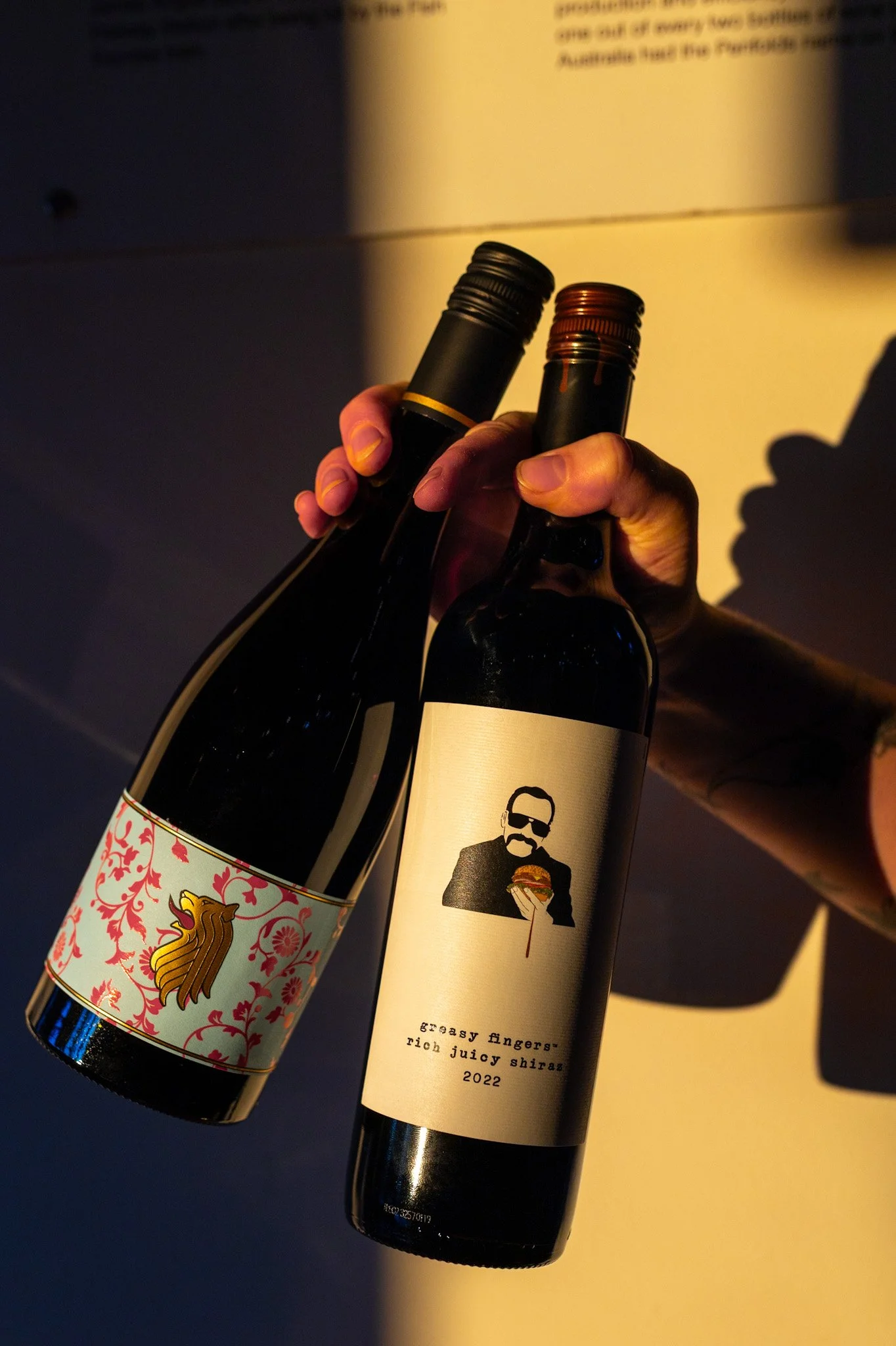 Two wine bottles being held up in front of a partly lit wall, one with a decorative label and the other with a label featuring a man in sunglasses holding a burger.