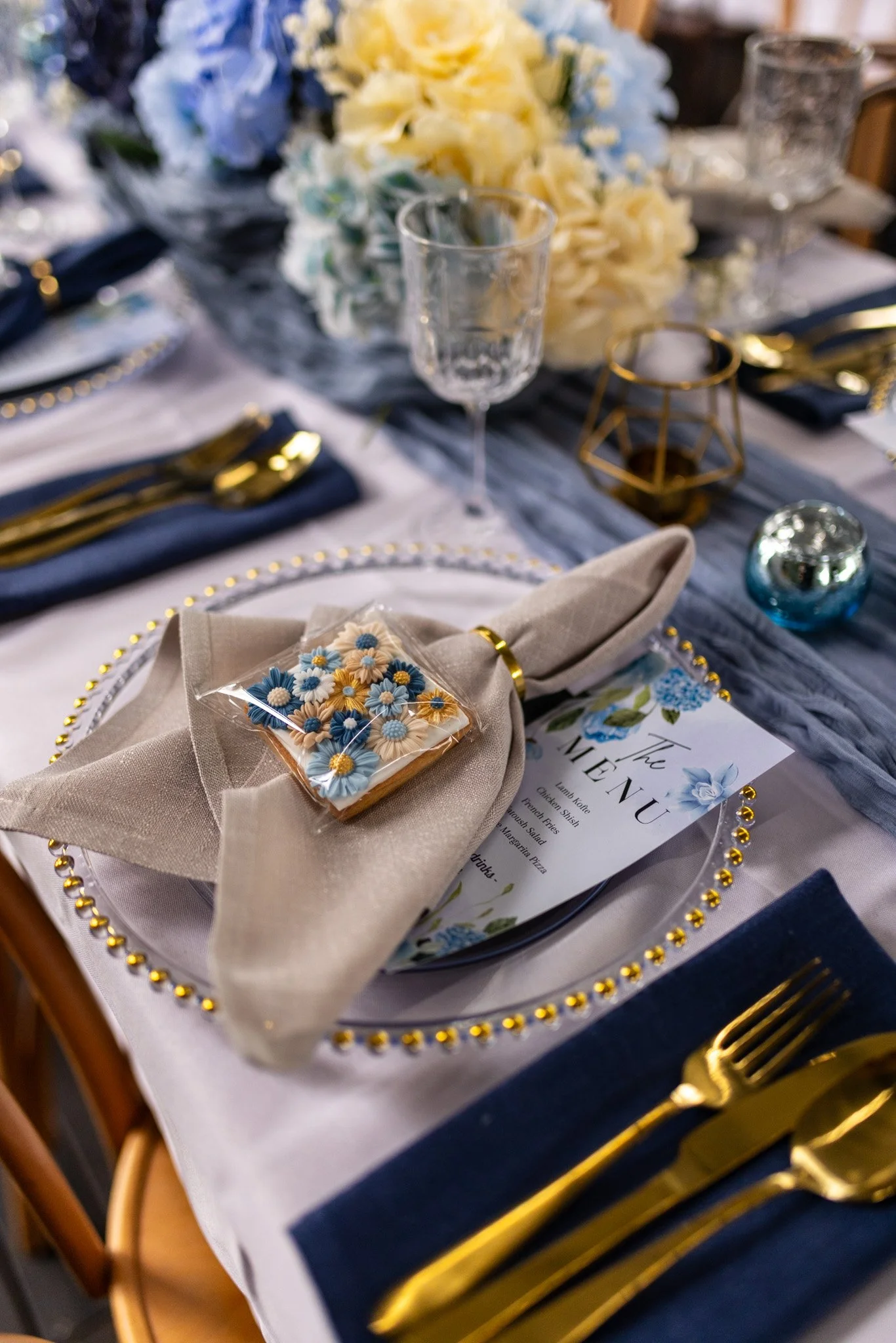 A formal dining table decorated with flowers, gold utensils, and a menu card, featuring a place setting with a napkin and decorated cookie.