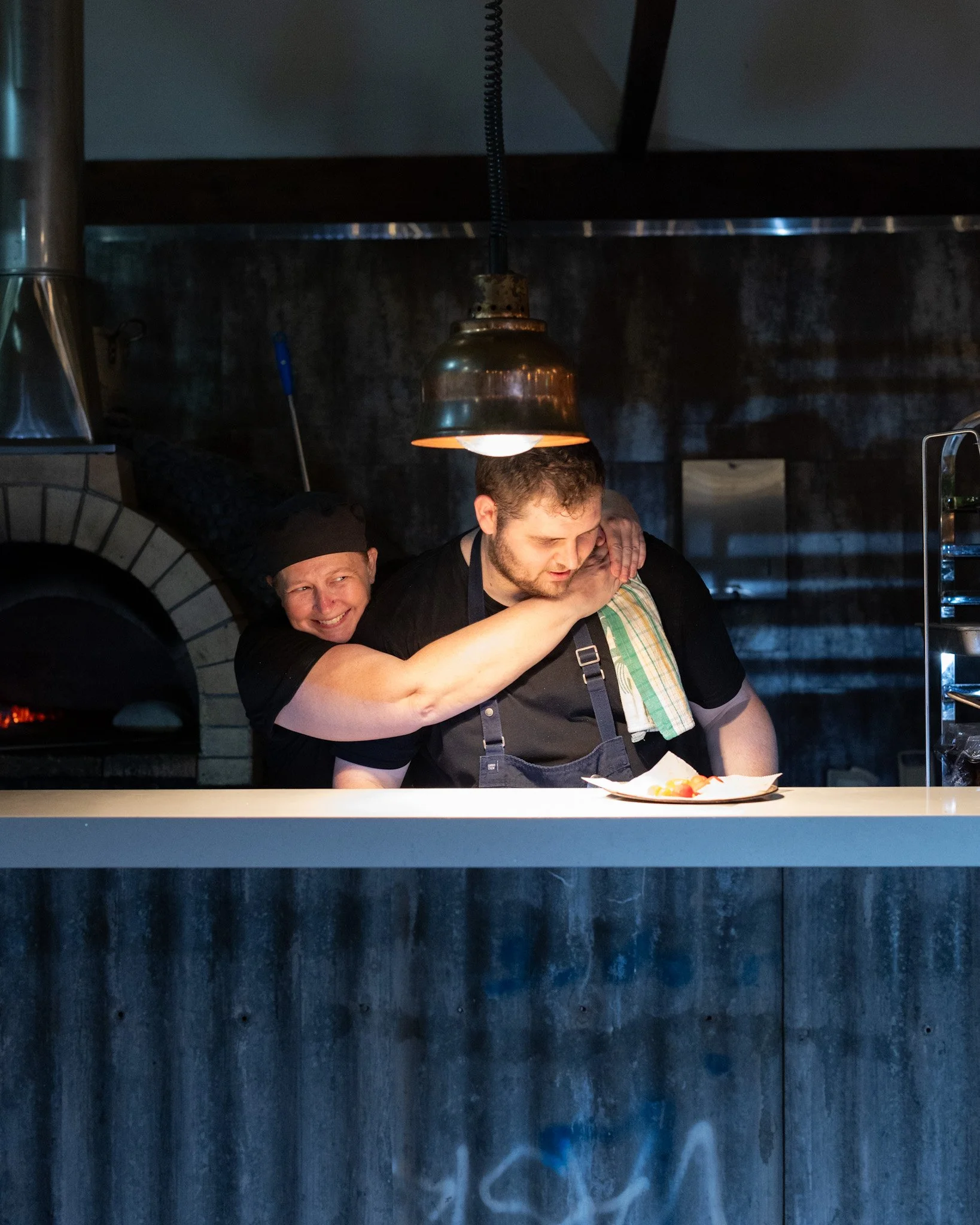A chef and a kitchen worker share a friendly hug behind a restaurant counter, with a pizza in front of them, under a hanging light.