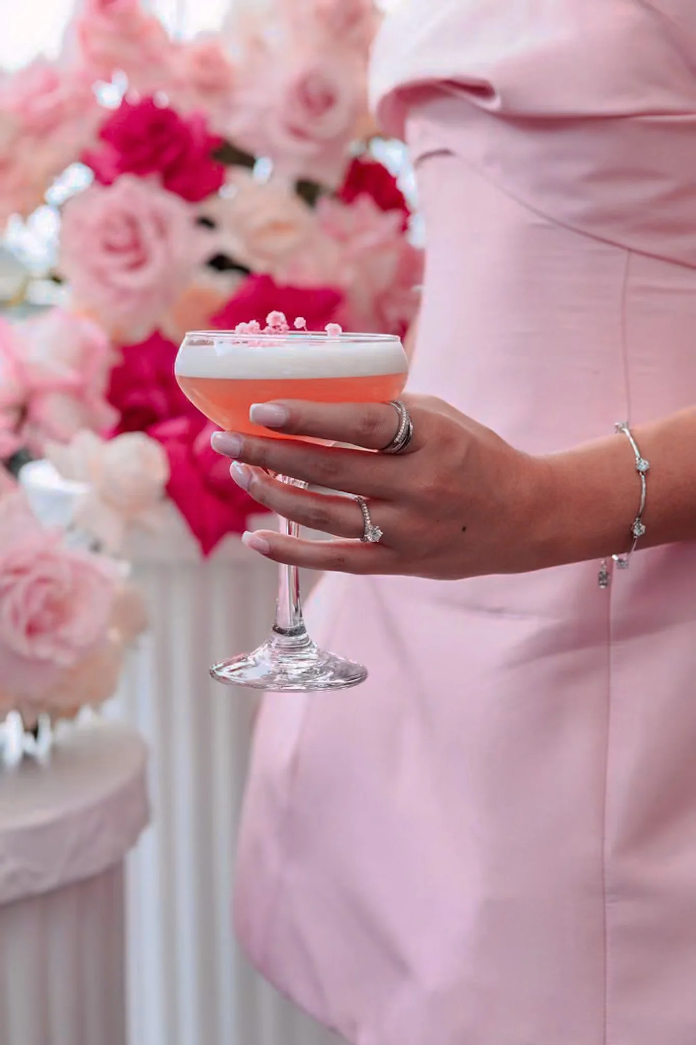 A woman in a pink dress holding a pink cocktail glass with white foam and pink sprinkles, standing in front of pink and white floral decorations.