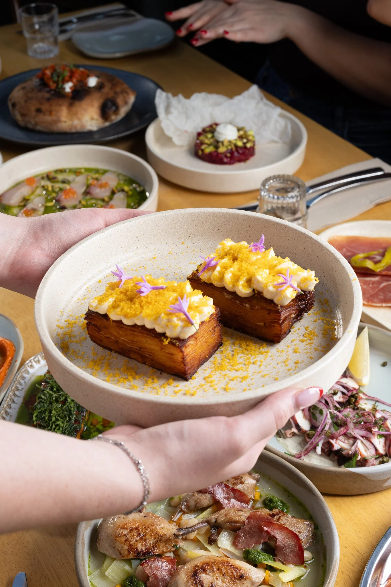 Person holding a plate with two pieces of dessert, topped with cream, yellow crumbs, and purple flowers, over a variety of other dishes on a dining table.