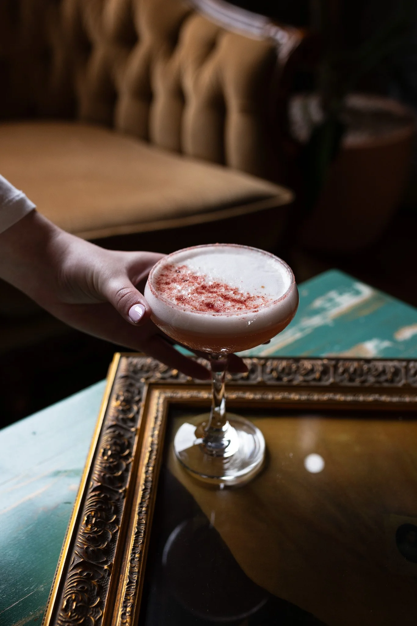 A person is holding a cocktail glass filled with a frothy drink topped with a sprinkle of ground spices, on a reflective surface with a decorative border, against a background of a tufted beige couch.