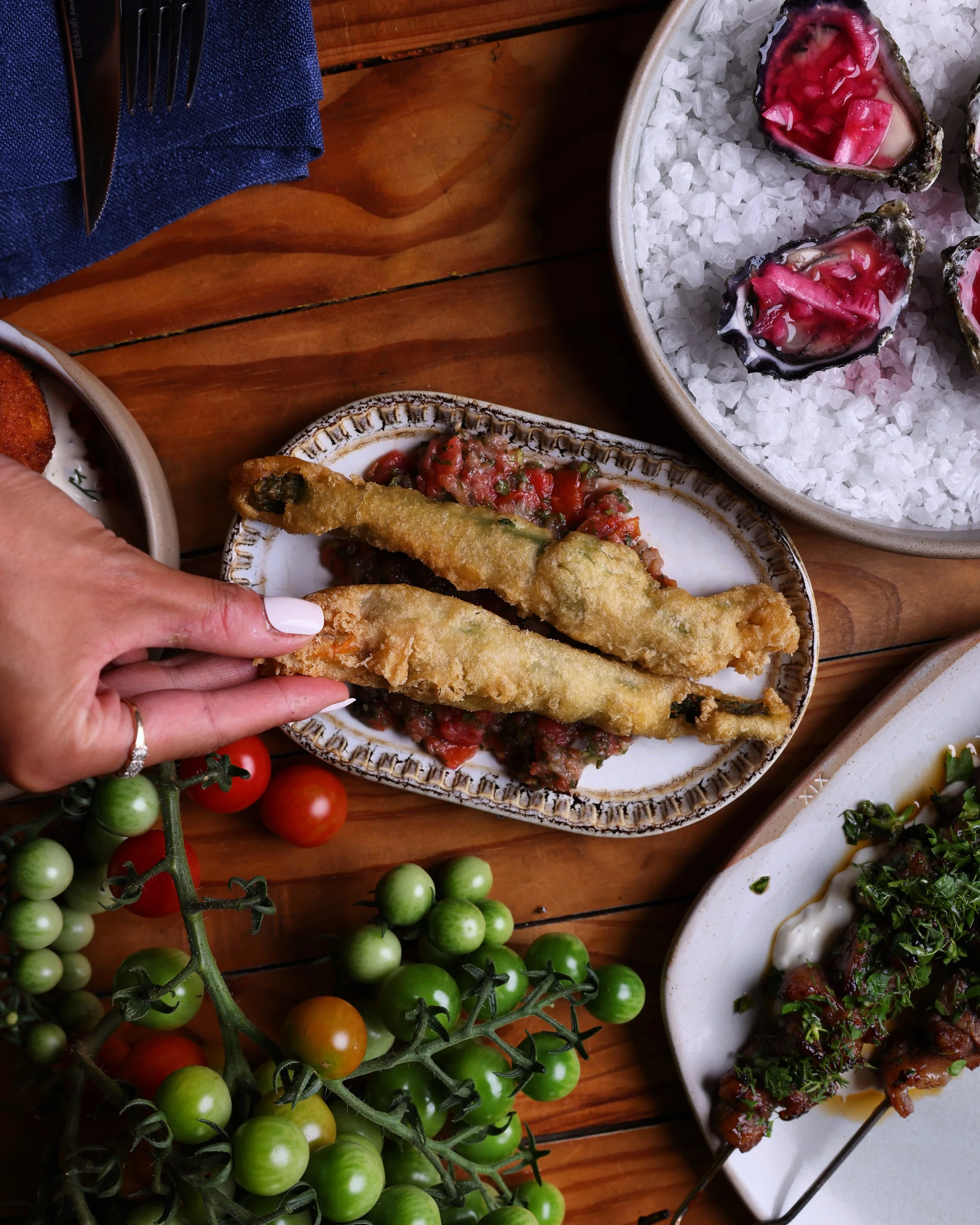 Fried fish fillets served on a plate with chopped tomatoes and herbs, a plate of oysters on ice with pink toppings, cherry tomatoes on a vine, and grilled meat skewers garnished with herbs on a white dish, all on a wooden table.